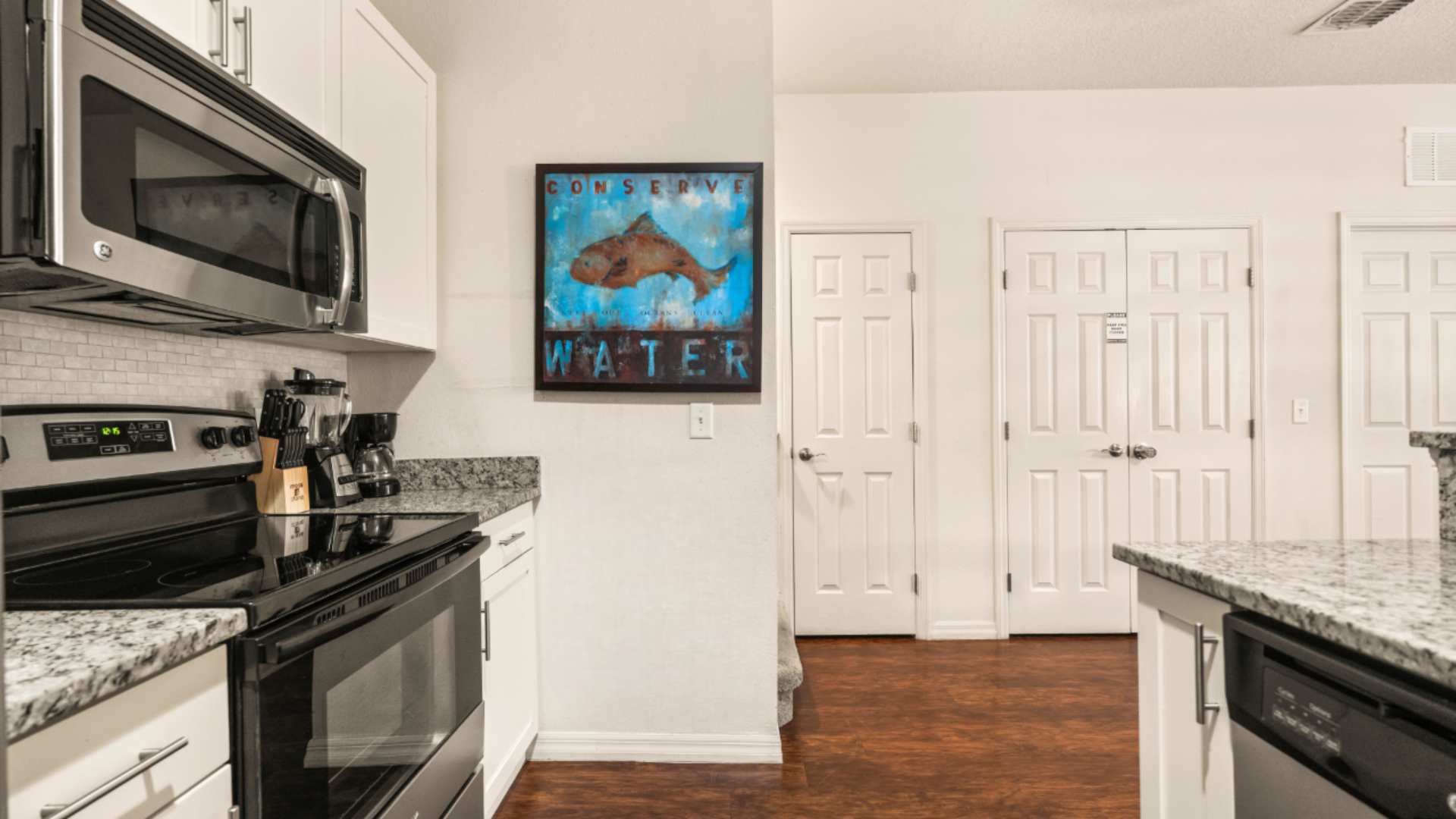 Modern kitchen featuring stainless steel appliances including a microwave and electric range, white shaker cabinets, granite countertops, and a white subway tile backsplash. A coffee maker and blender are visible on the counter.