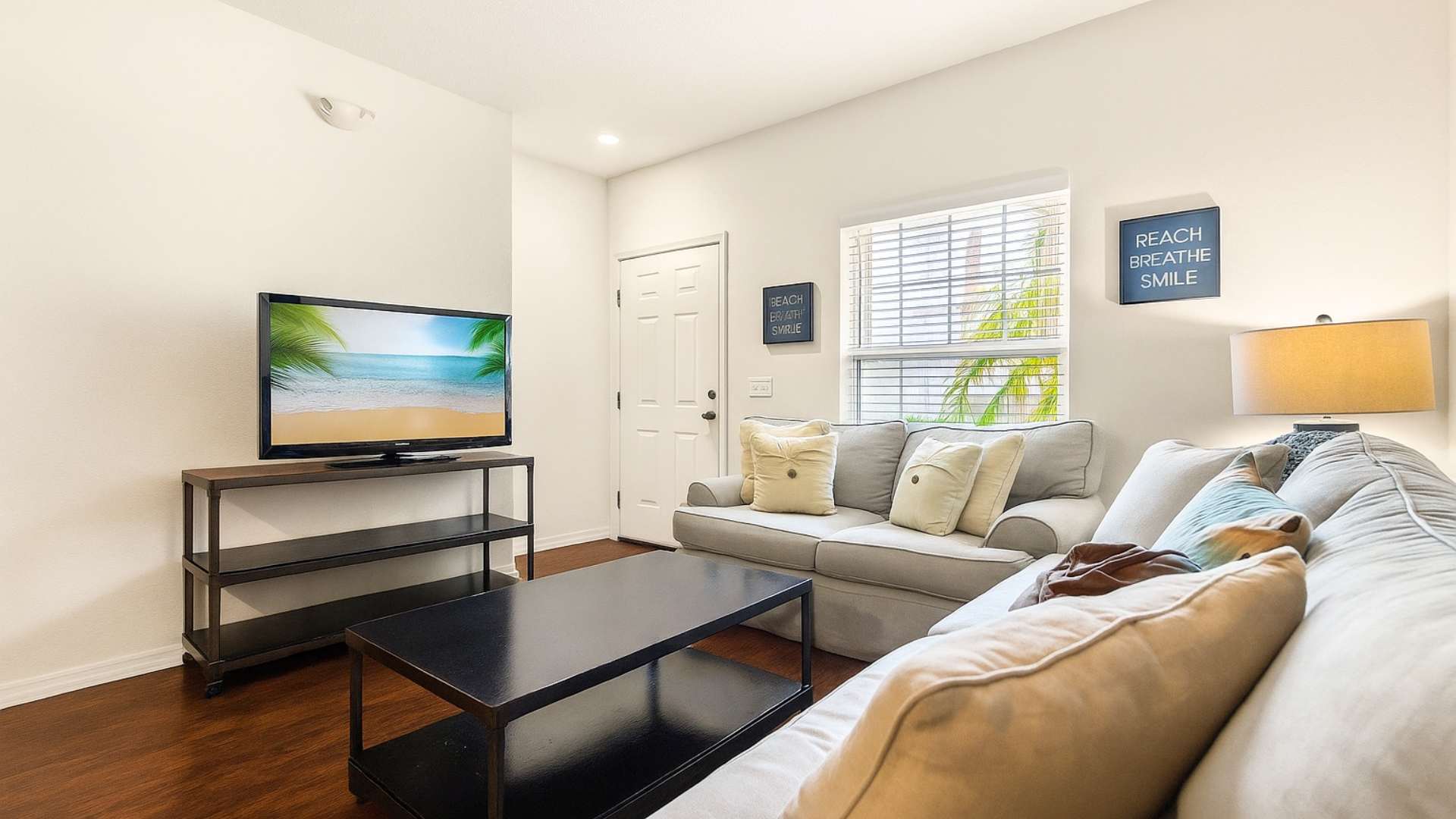 An inviting living room features dark hardwood floors, a comfortable light gray sofa, and a black coffee table. A flat-screen TV is set on a dark console, and a window with white blinds provides ample natural light to the space.