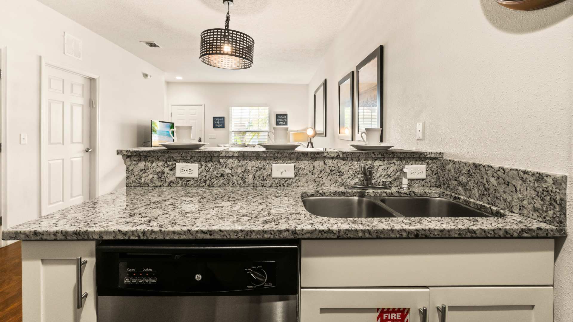Close-up view of the kitchen sink area with a double basin stainless steel sink set into granite countertops, and a stainless steel dishwasher below. White shaker cabinets and a modern pendant light are also visible.