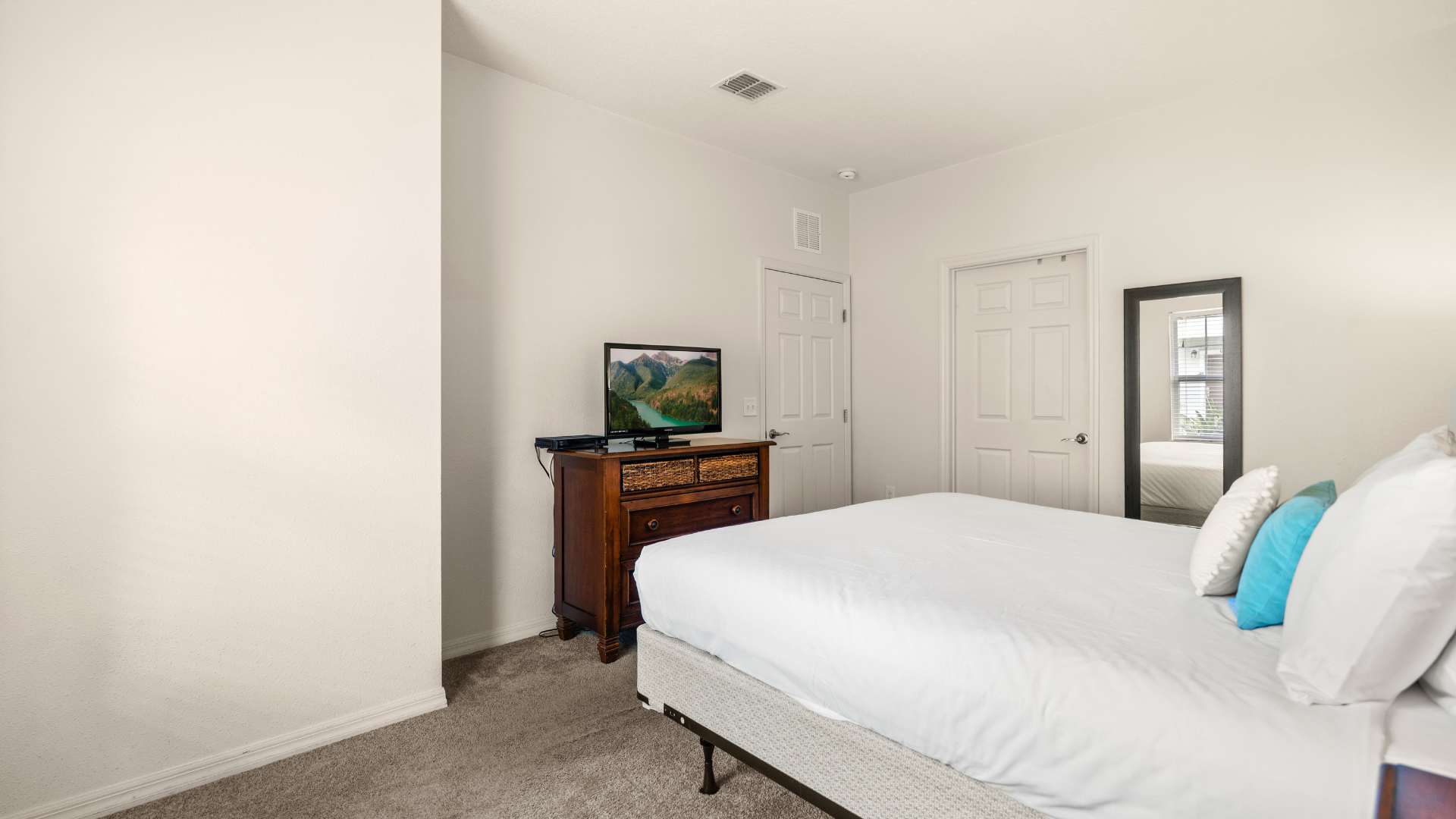 Bright bedroom featuring a king-sized bed with white bedding, a dark wood dresser with a flat-screen TV, and a full-length mirror leaning against the wall. The room has light gray carpet and white walls.
