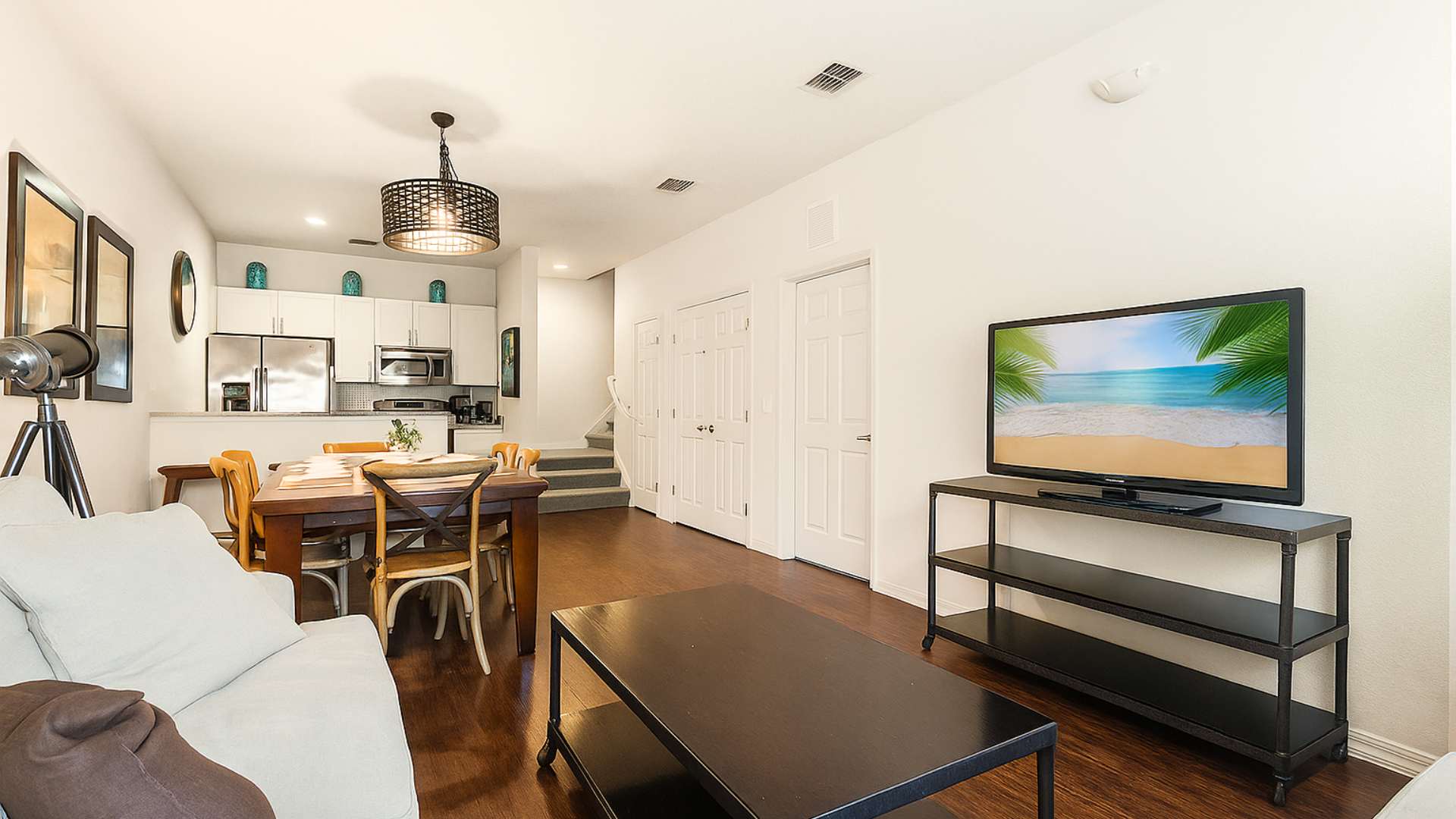 An expansive view captures the open-plan living, dining, and kitchen areas, all unified by dark hardwood floors. The space includes a light gray sofa, a wooden dining table, and a modern kitchen with white cabinets and stainless steel appliances, with stairs visible in the background.