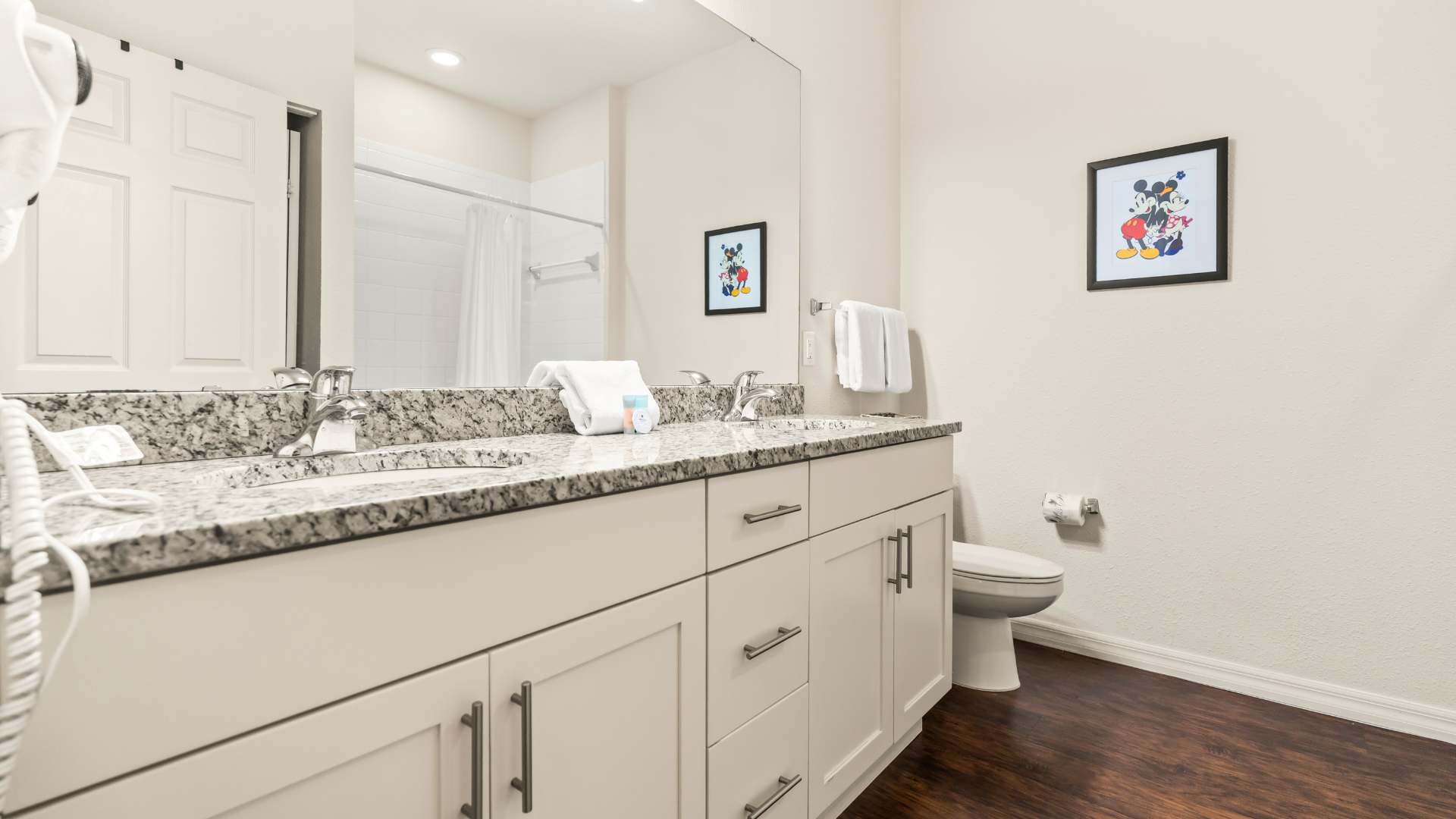 Modern bathroom with a double vanity featuring a speckled granite countertop, two sinks, and a large mirror. A toilet is visible next to the vanity, and two framed Mickey Mouse artworks adorn the light-colored walls.