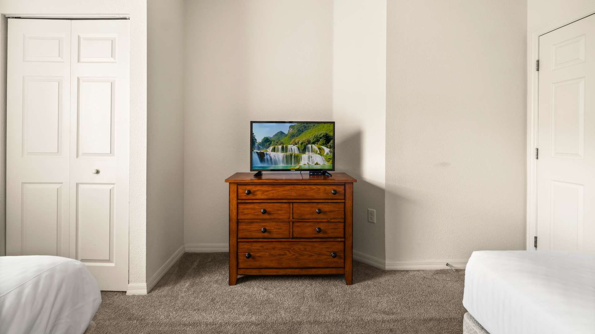 View of a bedroom corner with a wooden dresser topped by a television displaying a waterfall scene, flanked by two white closet doors. The room has light-colored walls and a grey carpeted floor.