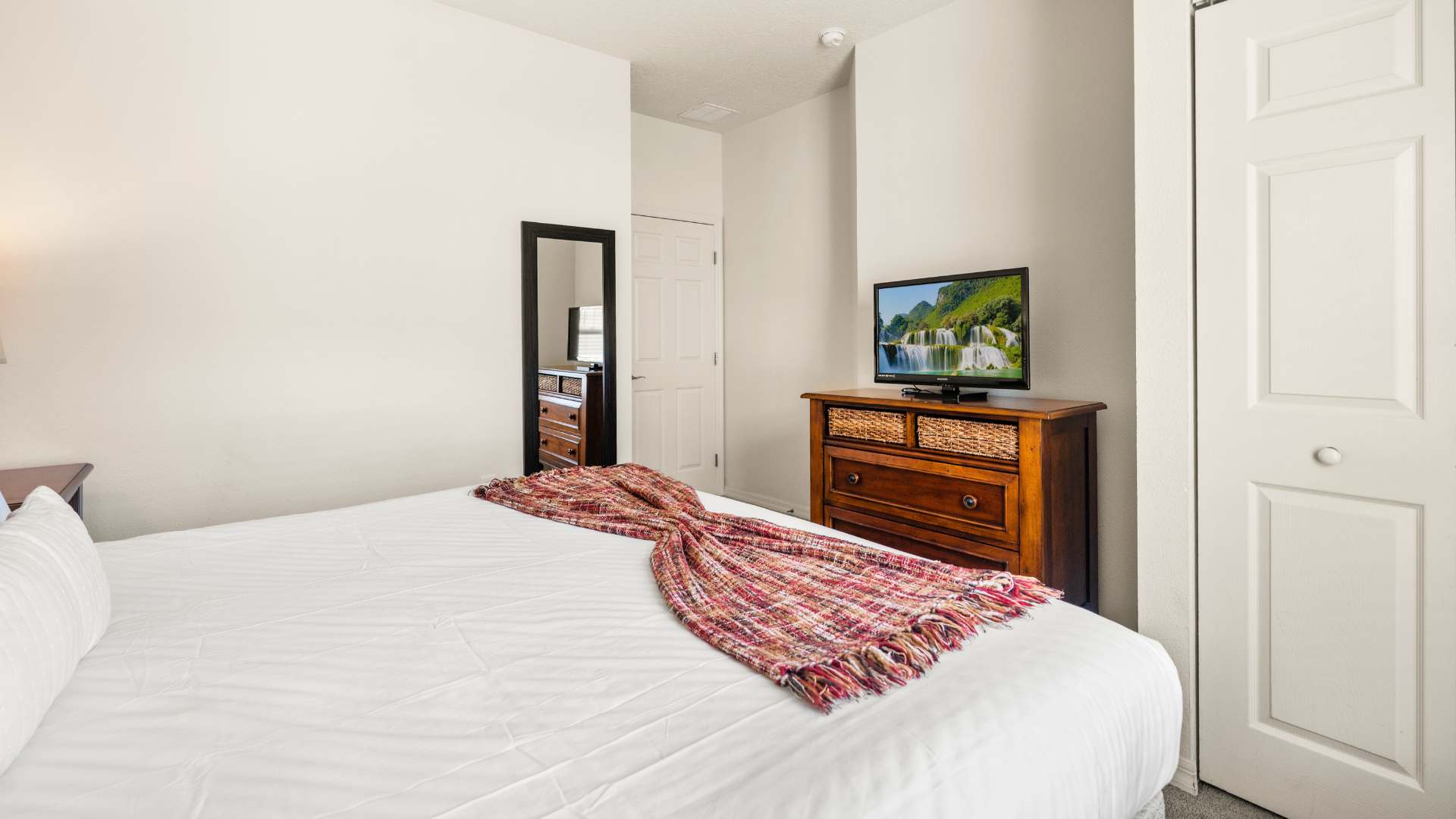 Bright bedroom featuring a large bed with white linens and a red plaid throw, a wooden dresser with a TV displaying a waterfall scene, and a full-length mirror reflecting a doorway.