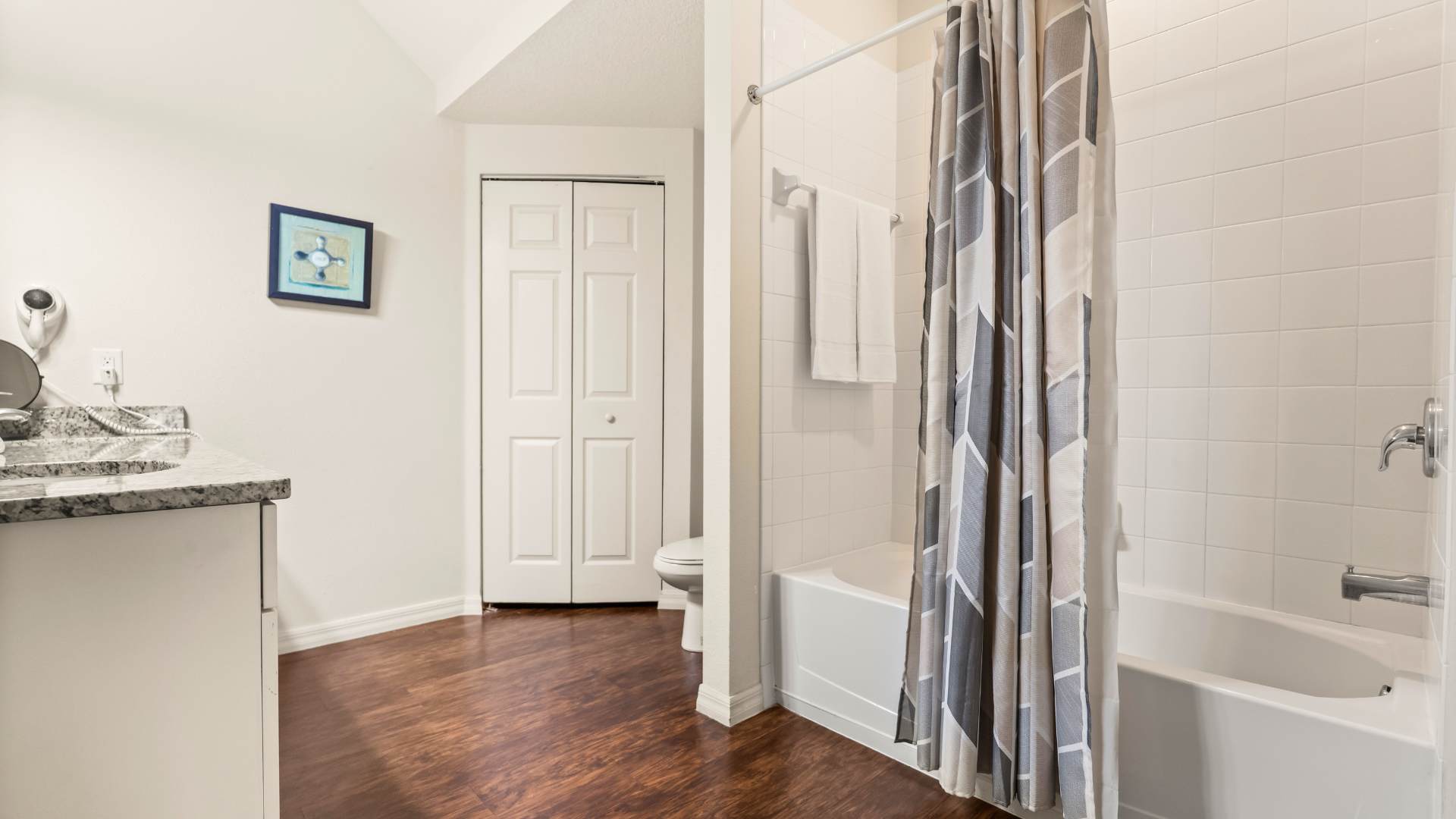 Bathroom with a white bathtub and shower combo, enclosed by a geometric pattern shower curtain, and white tiled walls. A toilet is visible next to a closet with bi-fold doors, and the room has dark wood-look flooring.
