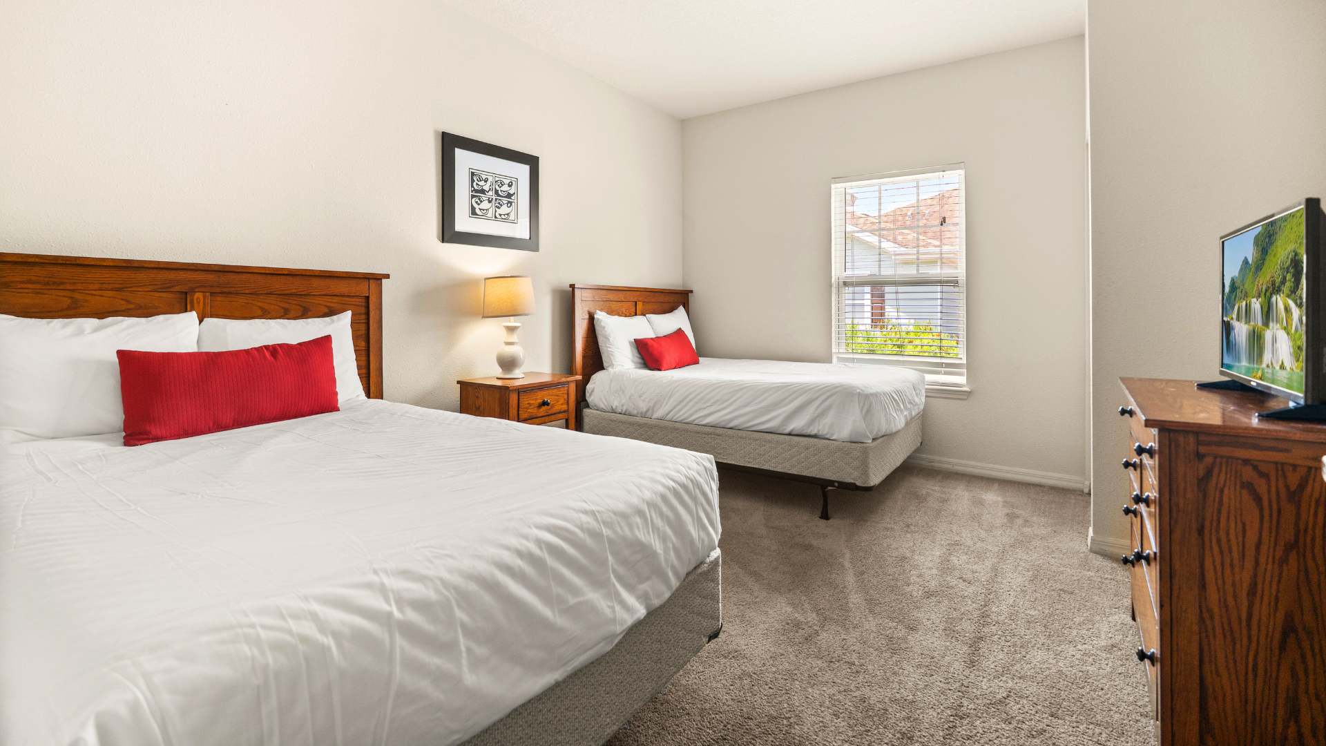 Cozy bedroom with two beds, one larger and one smaller, both with wooden headboards and red accent pillows. A wooden nightstand with a lamp sits between them, and a dresser with a TV is visible near a window with blinds.