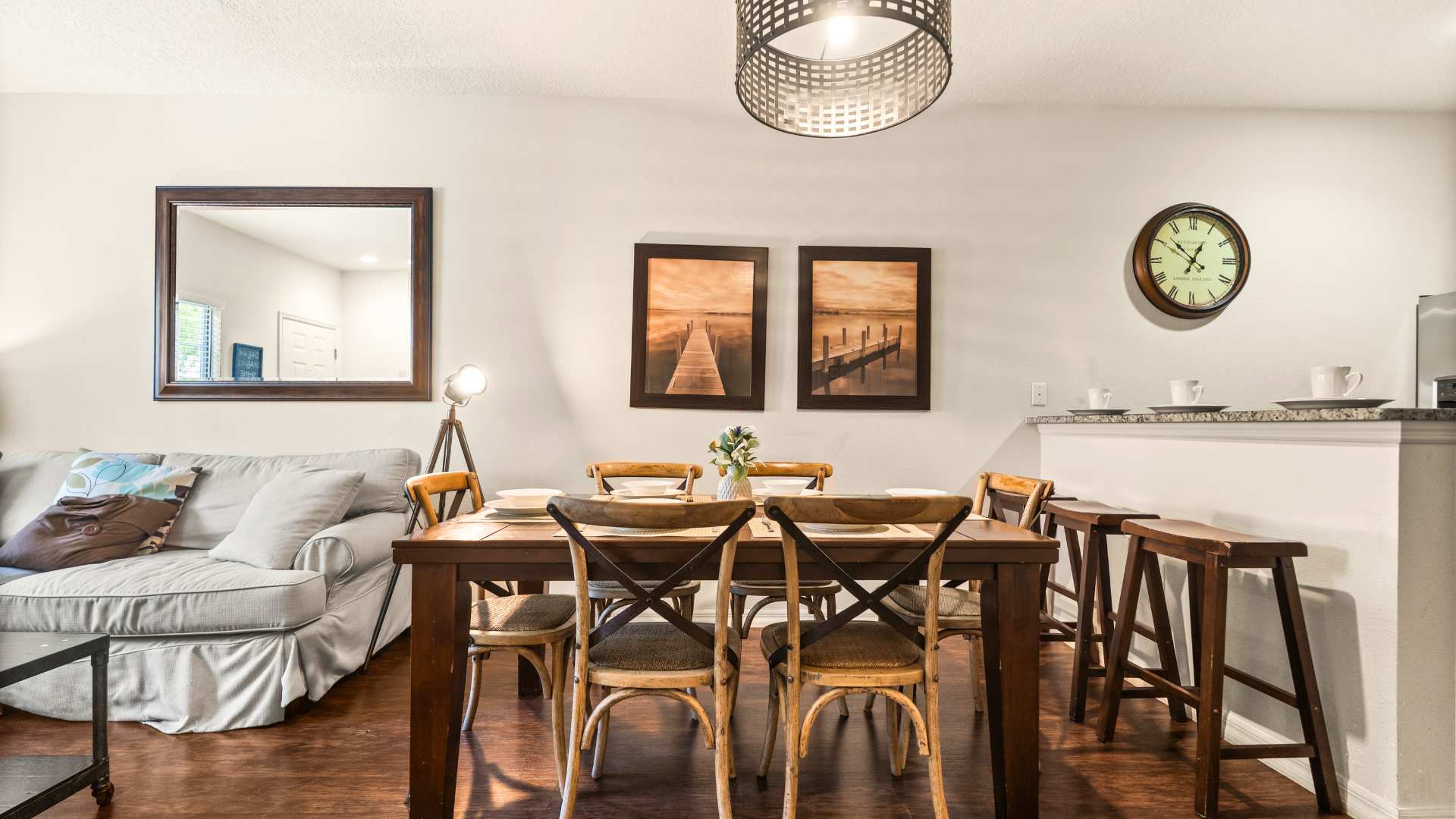 The dining area showcases a dark wooden table with six chairs, neatly set with white dishes and placemats, under a stylish caged chandelier. The adjacent living room, featuring a gray sofa and a large decorative mirror, is visible to the left.