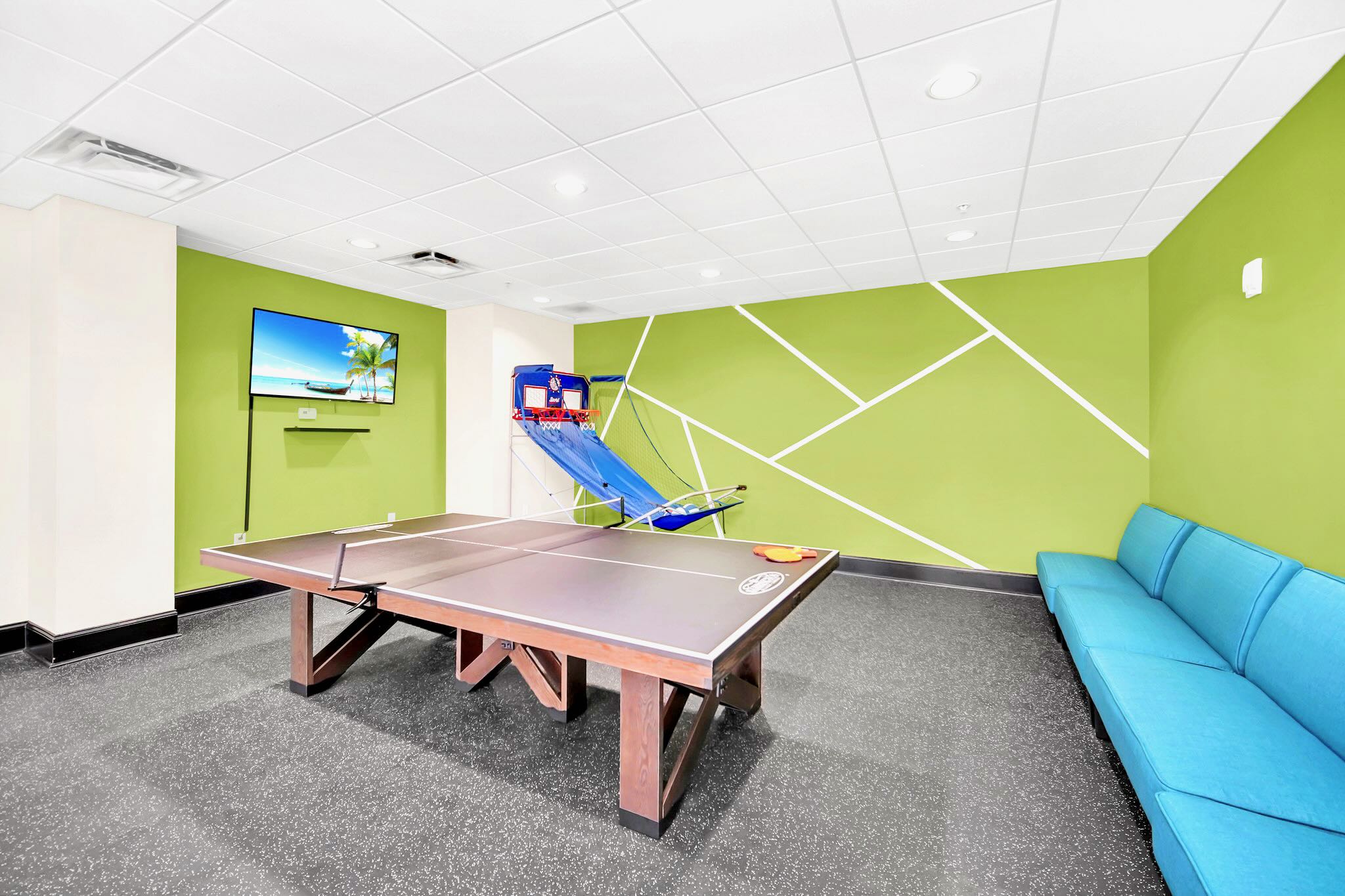 A vibrant game room with a ping-pong table at the center and a basketball arcade game in the corner. The room features a bright green accent wall with white geometric lines, a wall-mounted TV, and a blue sofa.