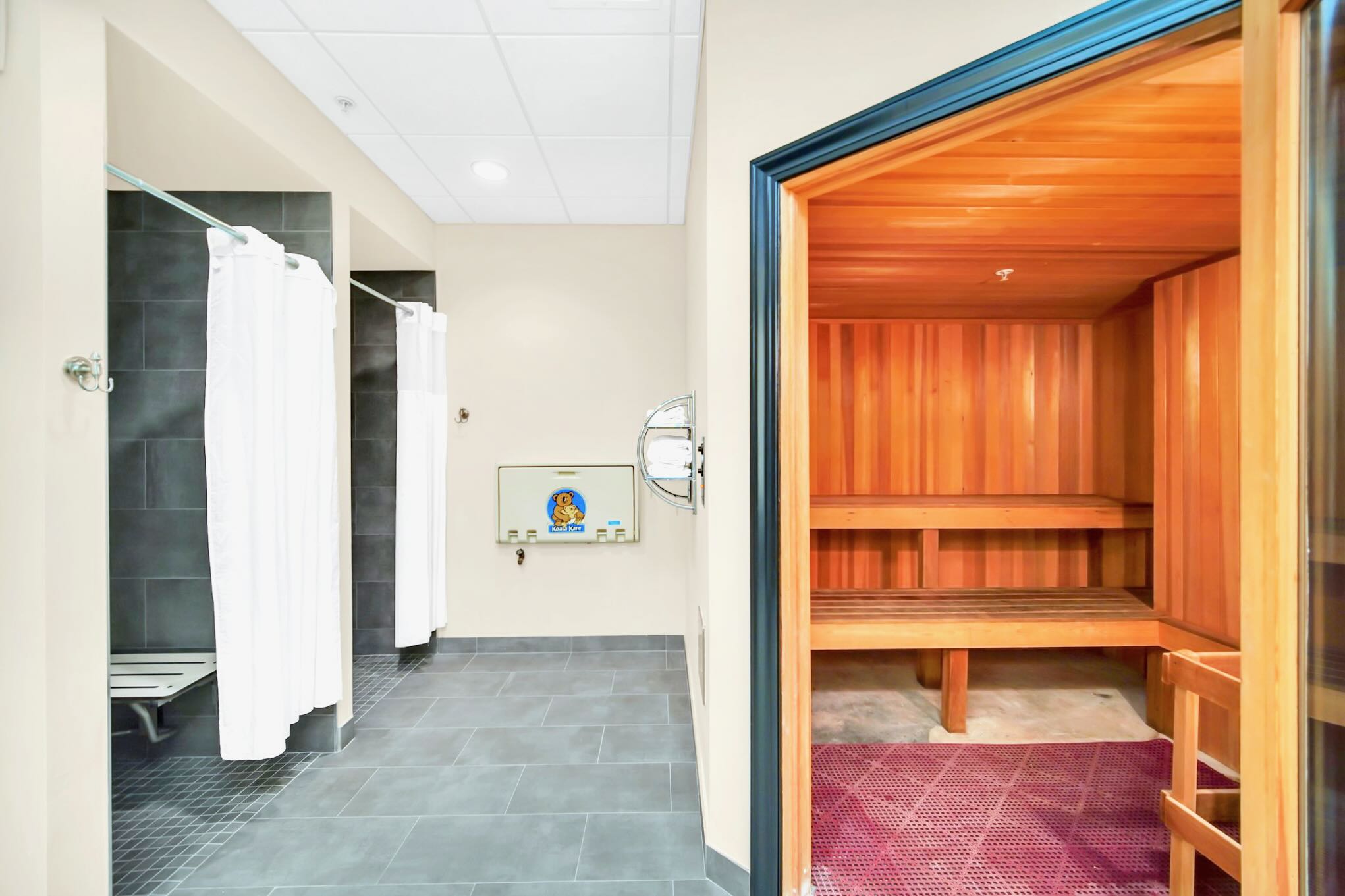 A clean and modern changing area featuring a wooden sauna with tiered benches on the right, and two shower stalls with white curtains and dark tiled walls on the left. A baby changing station is mounted on the wall.