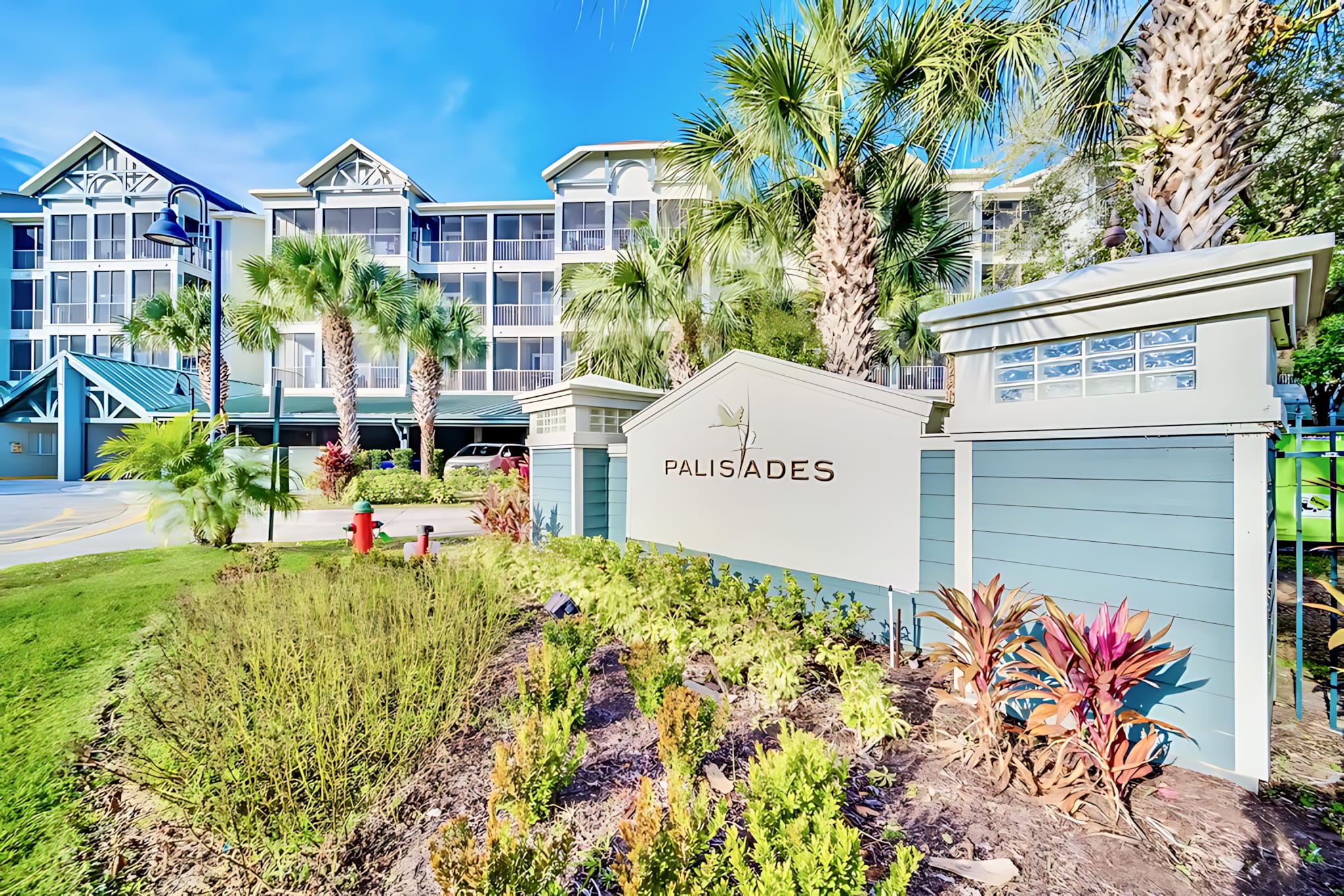 A vibrant exterior shot featuring the 'PALISADES' entrance sign with lush green landscaping, set against a multi-story condominium building with blue siding and palm trees under a clear blue sky.