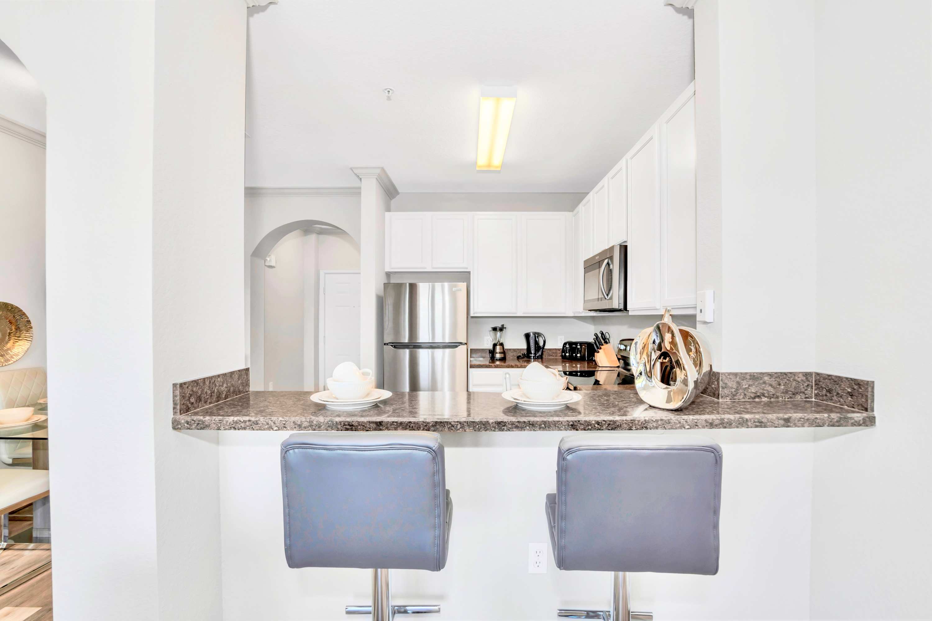 Modern kitchen with white shaker cabinets, dark granite countertops, stainless steel appliances, and a breakfast bar with two gray bar stools.