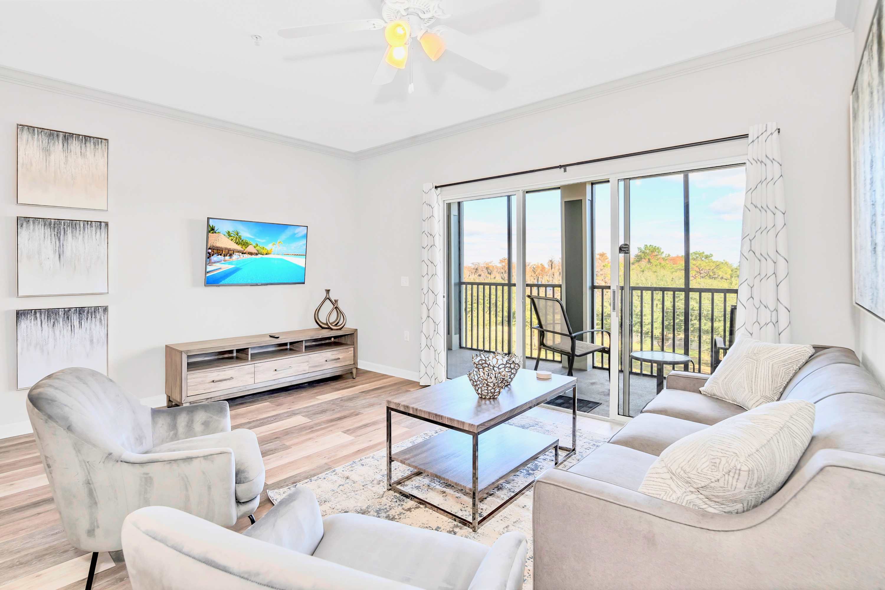 Bright living room with light wood-look flooring, a gray sofa, two light gray armchairs, and a dark wood coffee table on a patterned rug. A large TV is mounted on the left wall, and sliding glass doors open to a balcony with outdoor seating.