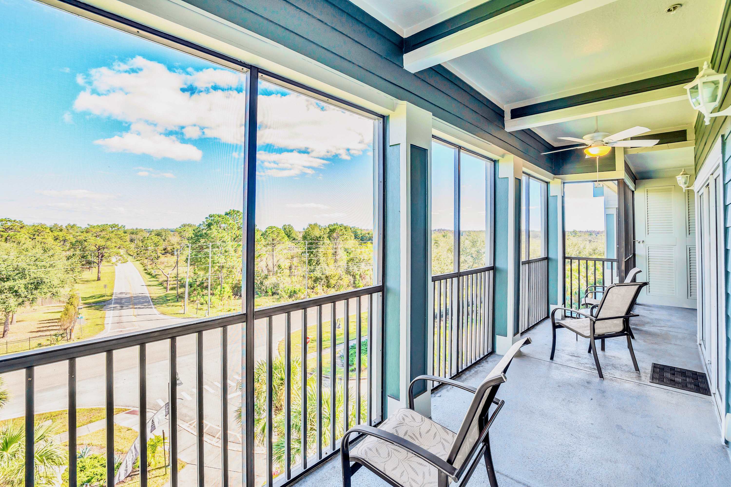 Spacious screened-in balcony with several outdoor chairs, offering expansive views of a tree-lined road and natural landscape. The balcony features a ceiling fan and a dark railing, providing a comfortable outdoor seating area.