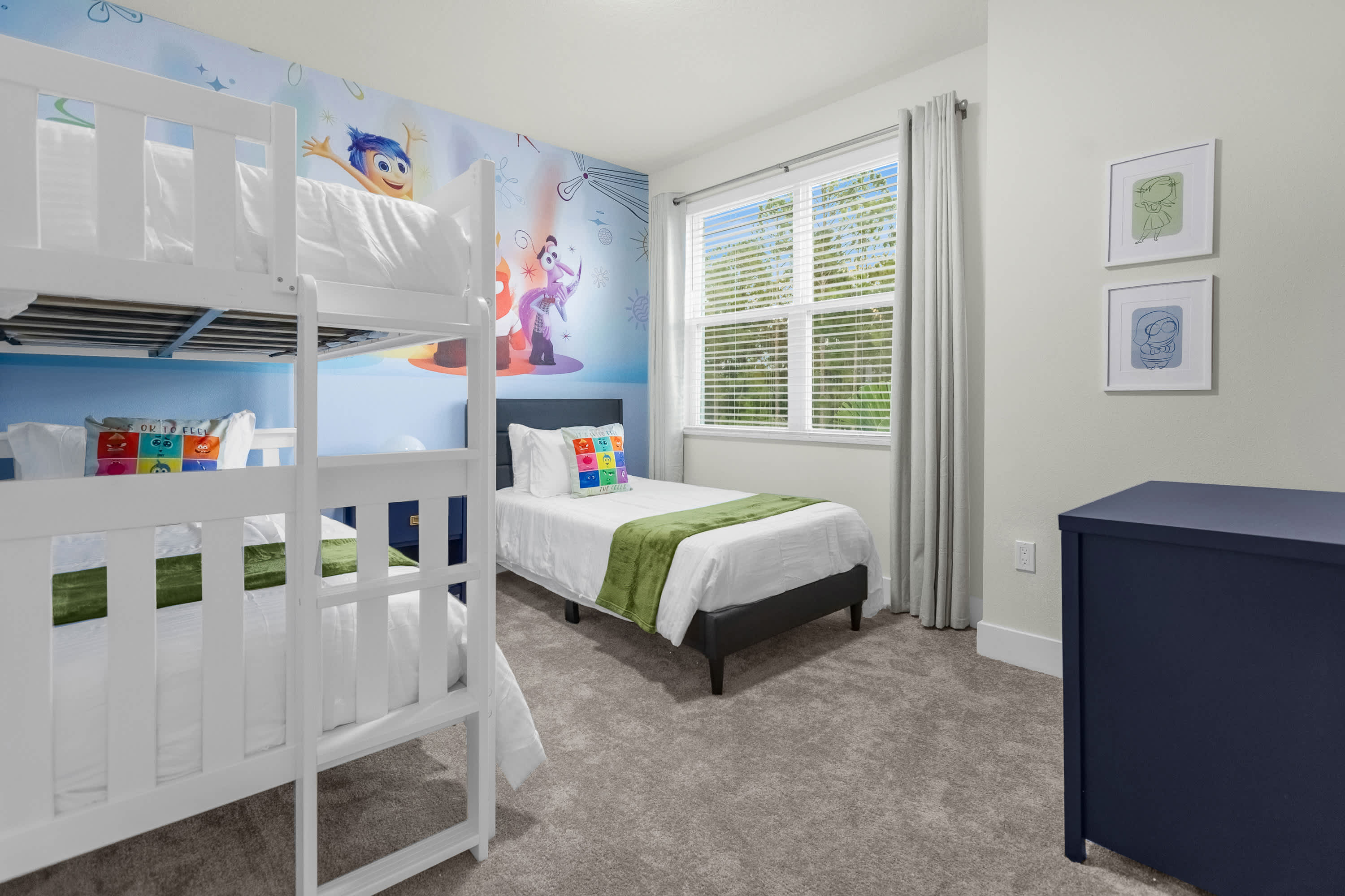 Another view of the vibrant kids' bedroom, showcasing the white wooden bunk bed and a single bed with a dark upholstered headboard, both dressed with green throws. The room features light blue walls, a colorful 'Inside Out' mural, a large window with blinds, and a dark blue dresser.