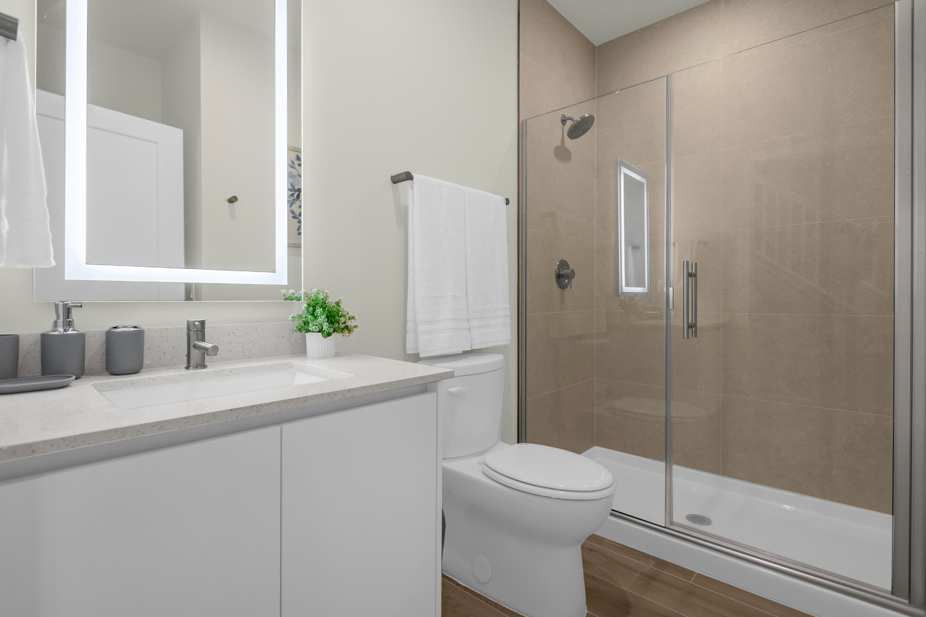 Modern bathroom featuring a white vanity with a light gray quartz countertop, a large illuminated mirror, and a glass-enclosed walk-in shower with light brown tiled walls. A white toilet is positioned next to the vanity, complemented by wood-look floor tiles.