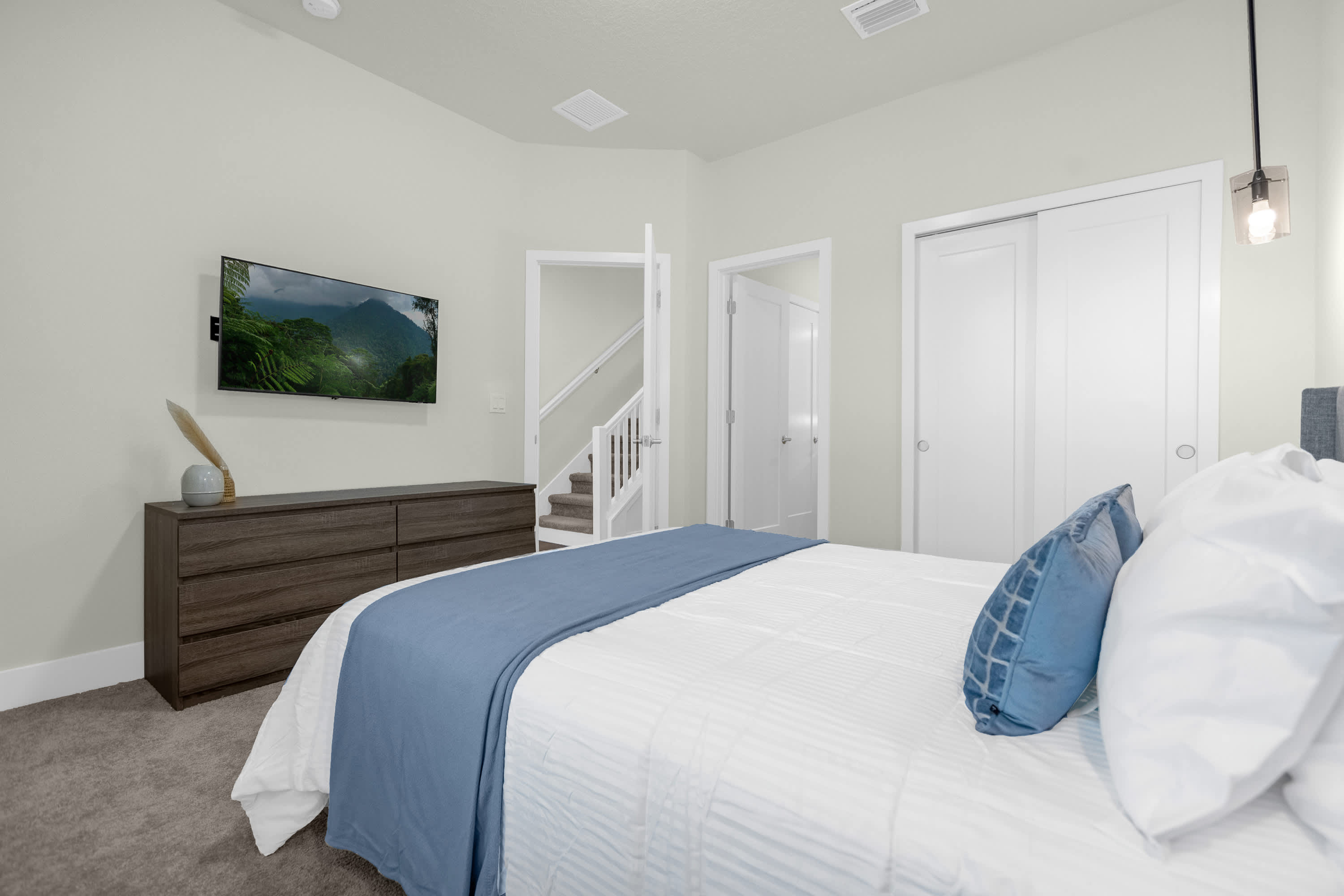 Another view of bedroom 2 showing a wall-mounted TV above a dark wood dresser, flanked by white sliding closet doors and a doorway revealing a staircase.