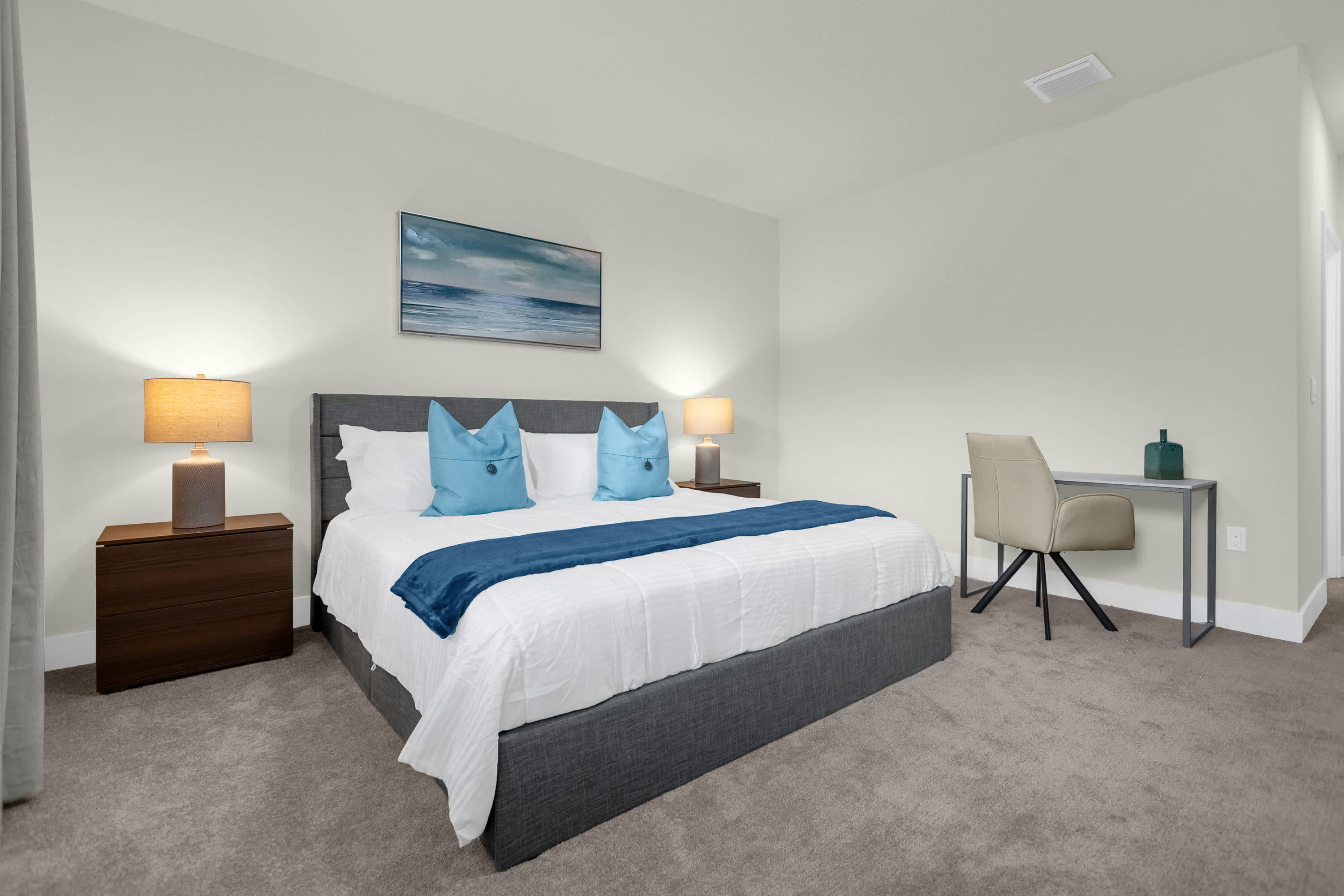 Spacious bedroom with a king-sized bed featuring a gray upholstered headboard, two dark wood nightstands with lamps, a modern desk with a chair, and light gray carpeted flooring.
