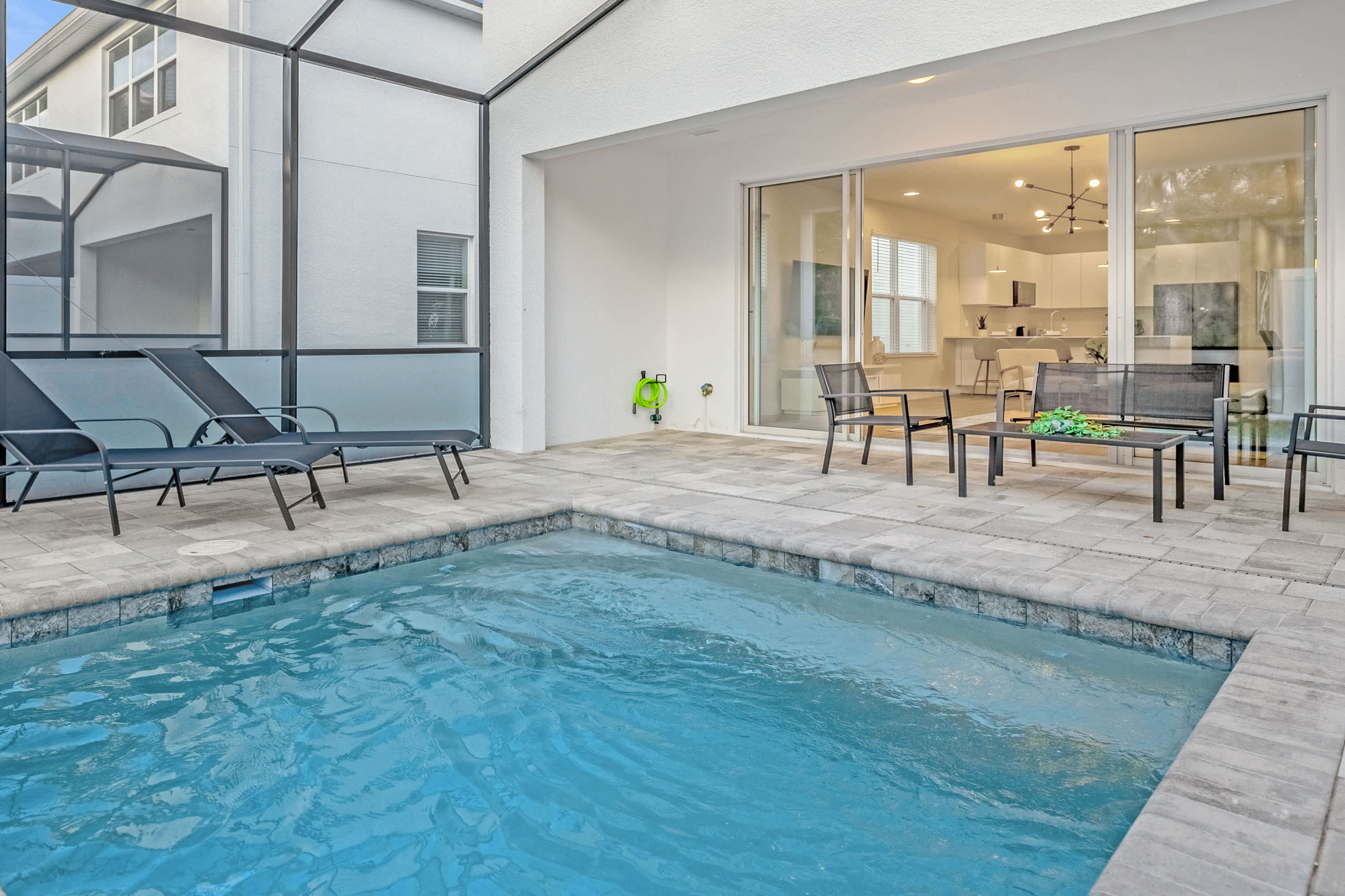 Outdoor screened patio area featuring a small rectangular private pool with light gray paver decking. Two black lounge chairs are positioned by the pool, with a separate seating area including a table and chairs visible further back.