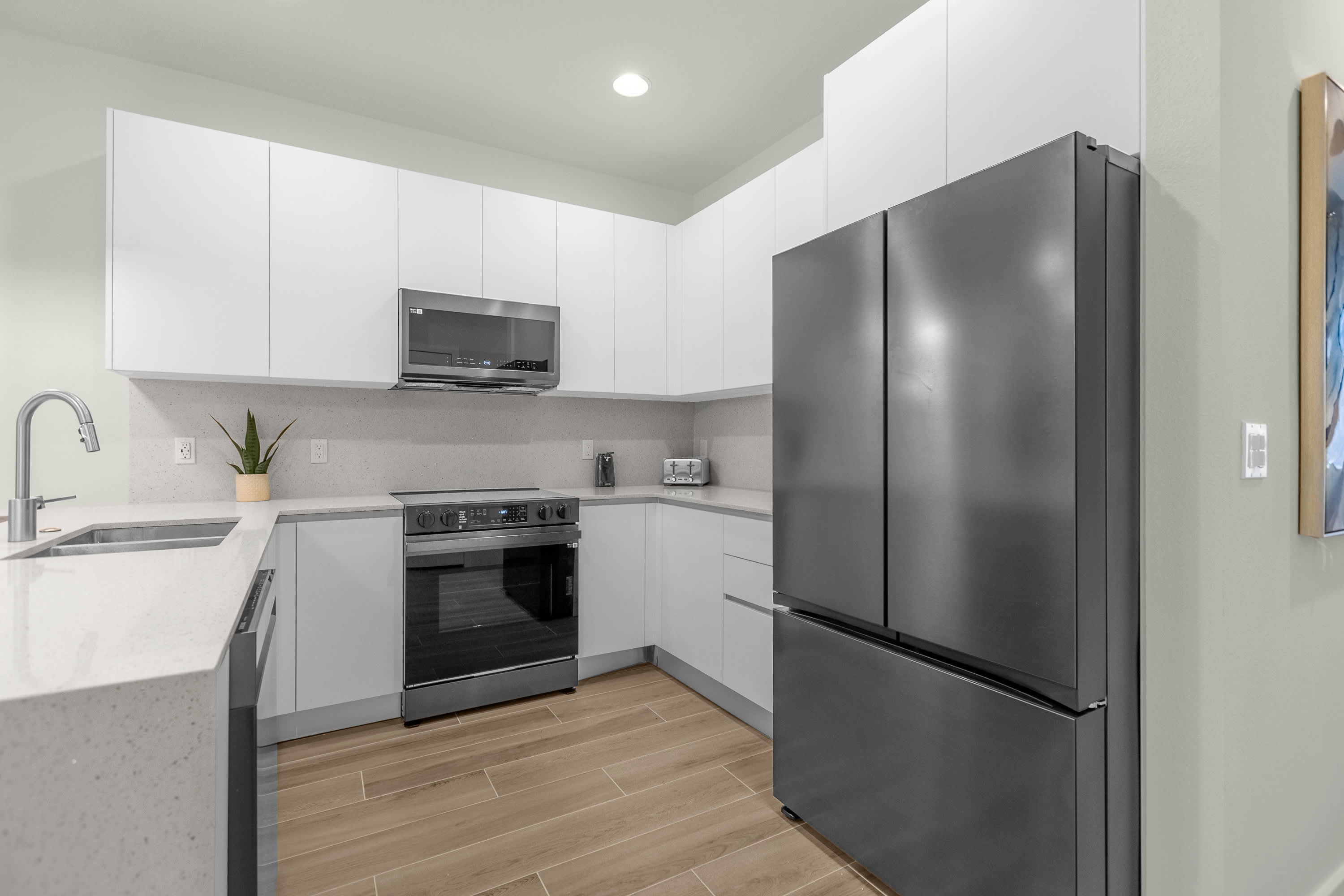 Modern kitchen with white flat-panel cabinets, light quartz countertops, and dark stainless steel appliances including a French door refrigerator, oven, and microwave. The wood-look flooring extends throughout the space.