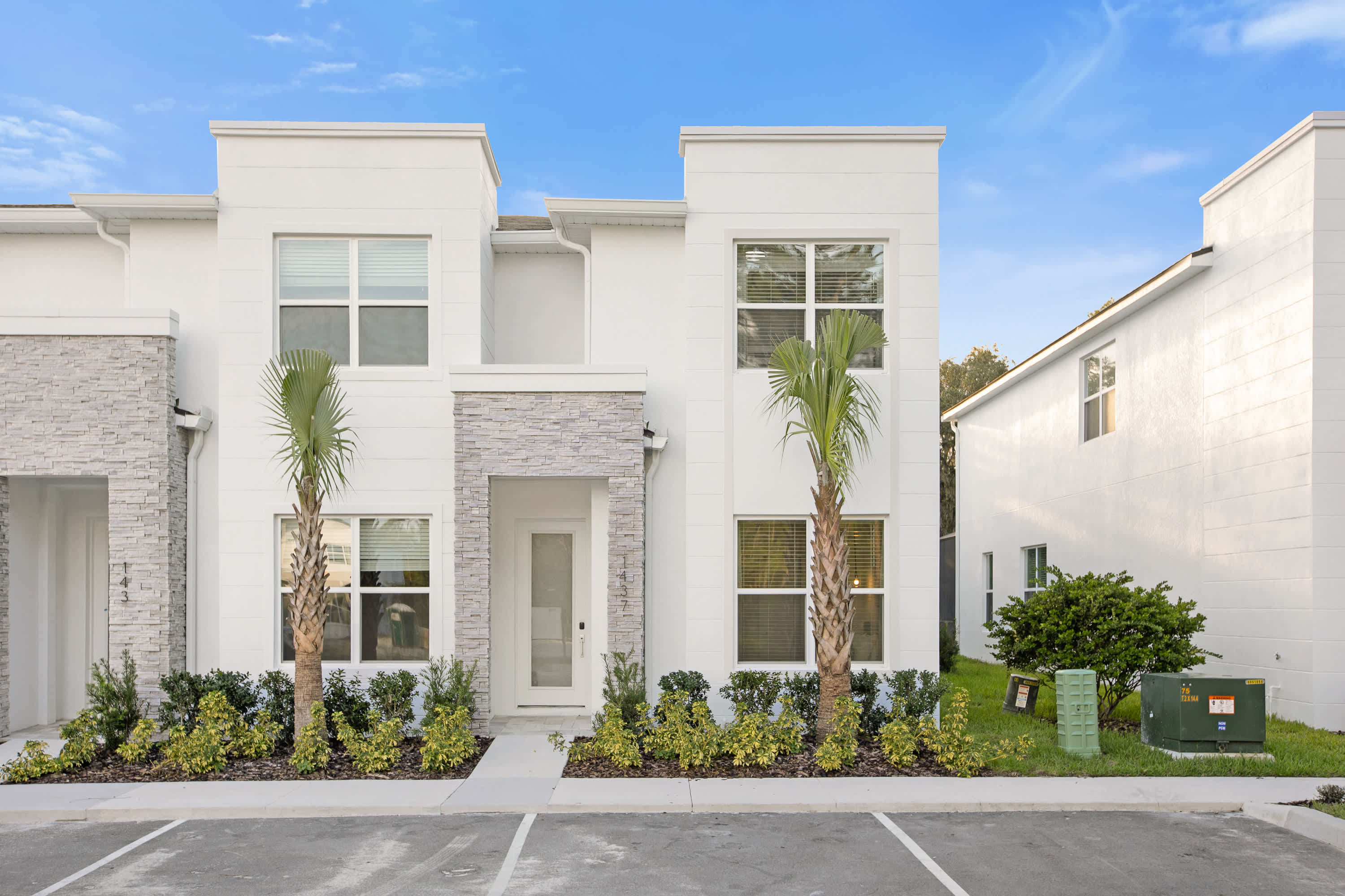 Modern white townhouse exterior with a clean, minimalist design, featuring stacked stone accents around the entrance and two palm trees flanking the front door. The property has multiple windows with white blinds and a small landscaped area with green shrubs.
