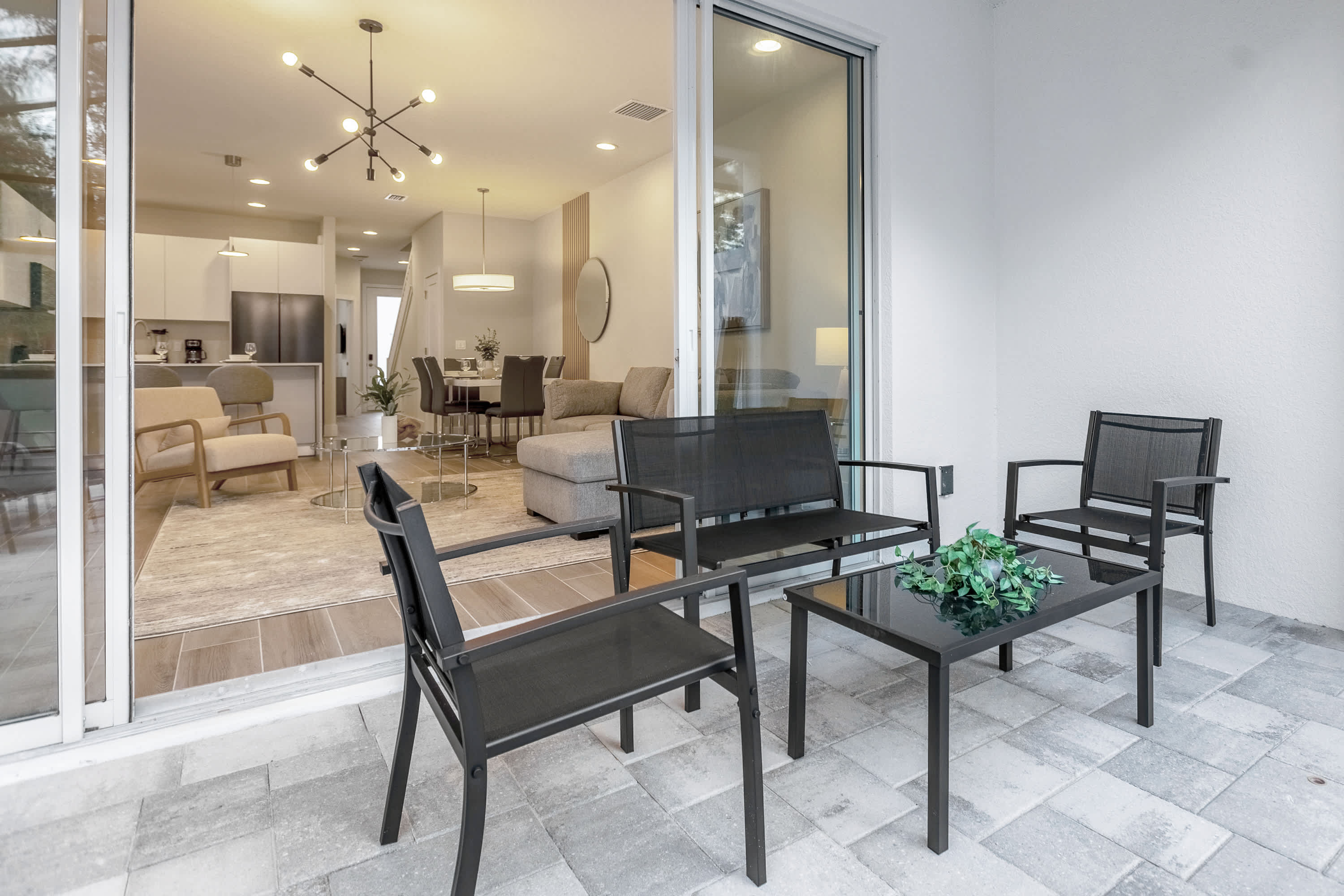 Close-up view of the outdoor patio seating area, featuring a black metal coffee table and two chairs, with a bench visible on the left. Sliding glass doors connect the patio to the interior living space, offering a glimpse of the modern decor.