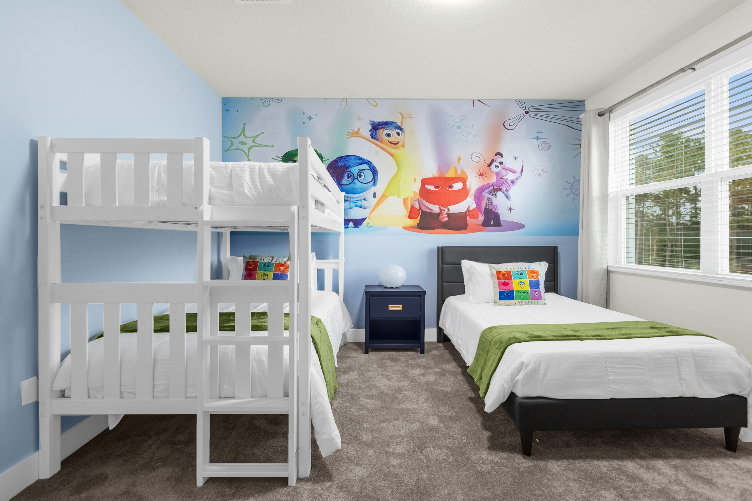 Vibrant kids' bedroom with light blue walls and a colorful mural featuring 'Inside Out' characters. It includes a white wooden bunk bed and a single bed with a dark upholstered headboard, both dressed with white linens and green throws, on a gray carpeted floor.
