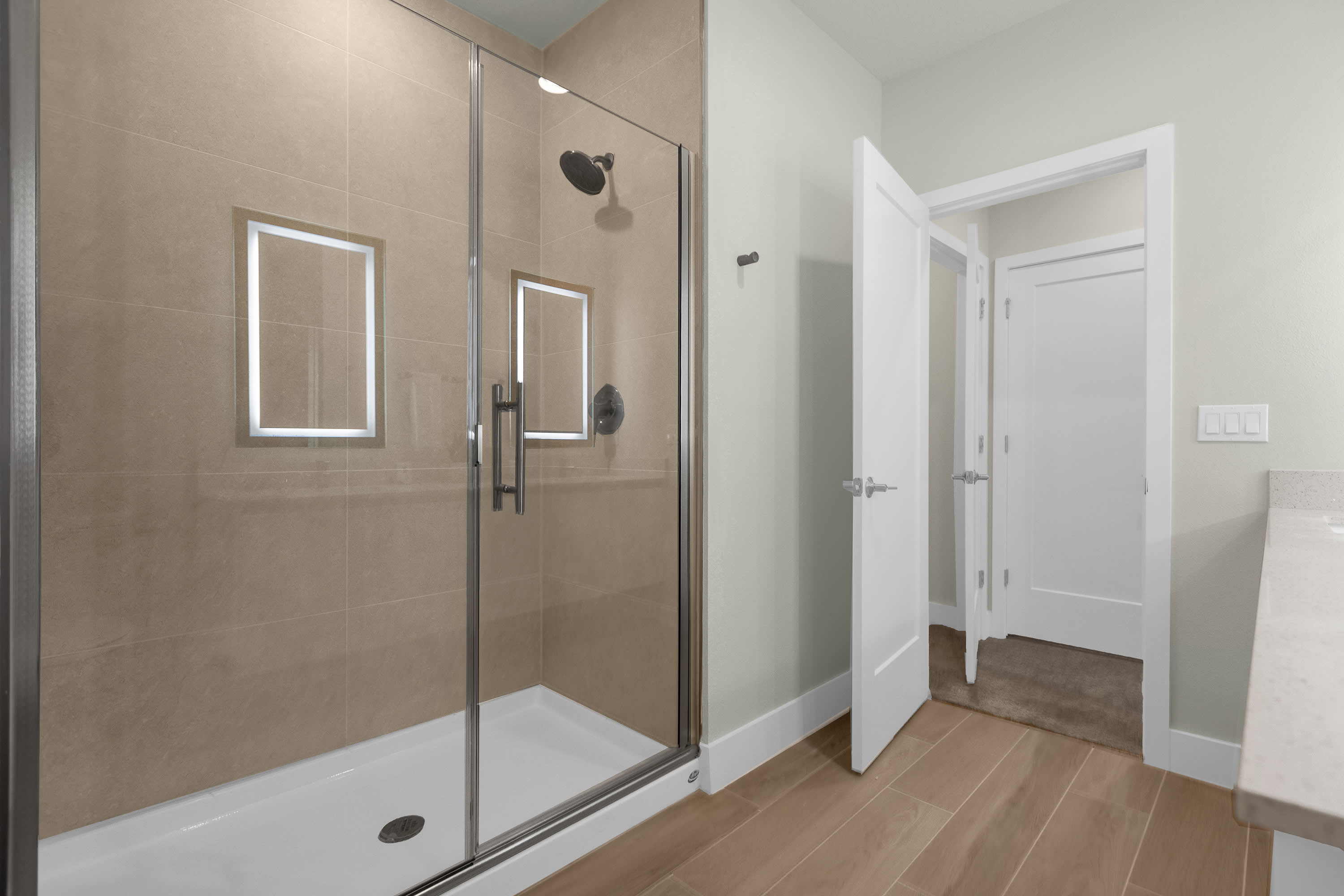 Contemporary walk-in shower with light brown tiled walls, a sleek glass enclosure, and a white shower pan, positioned next to a doorway leading to another room.