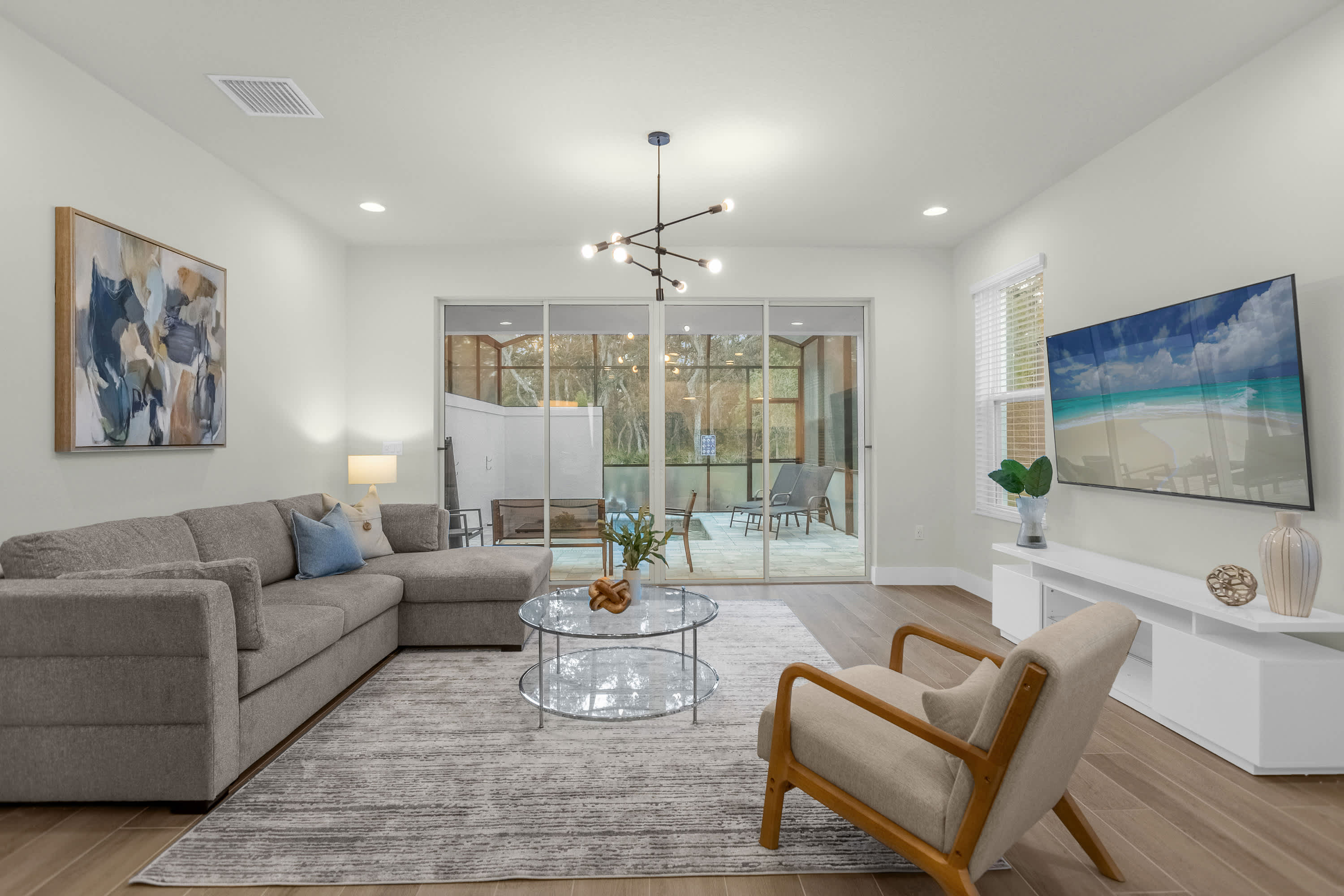 Bright living room with a large gray sectional sofa and a glass coffee table on a textured area rug. Sliding glass doors lead to an outdoor patio, and a white TV console with a wall-mounted TV is visible on the right.
