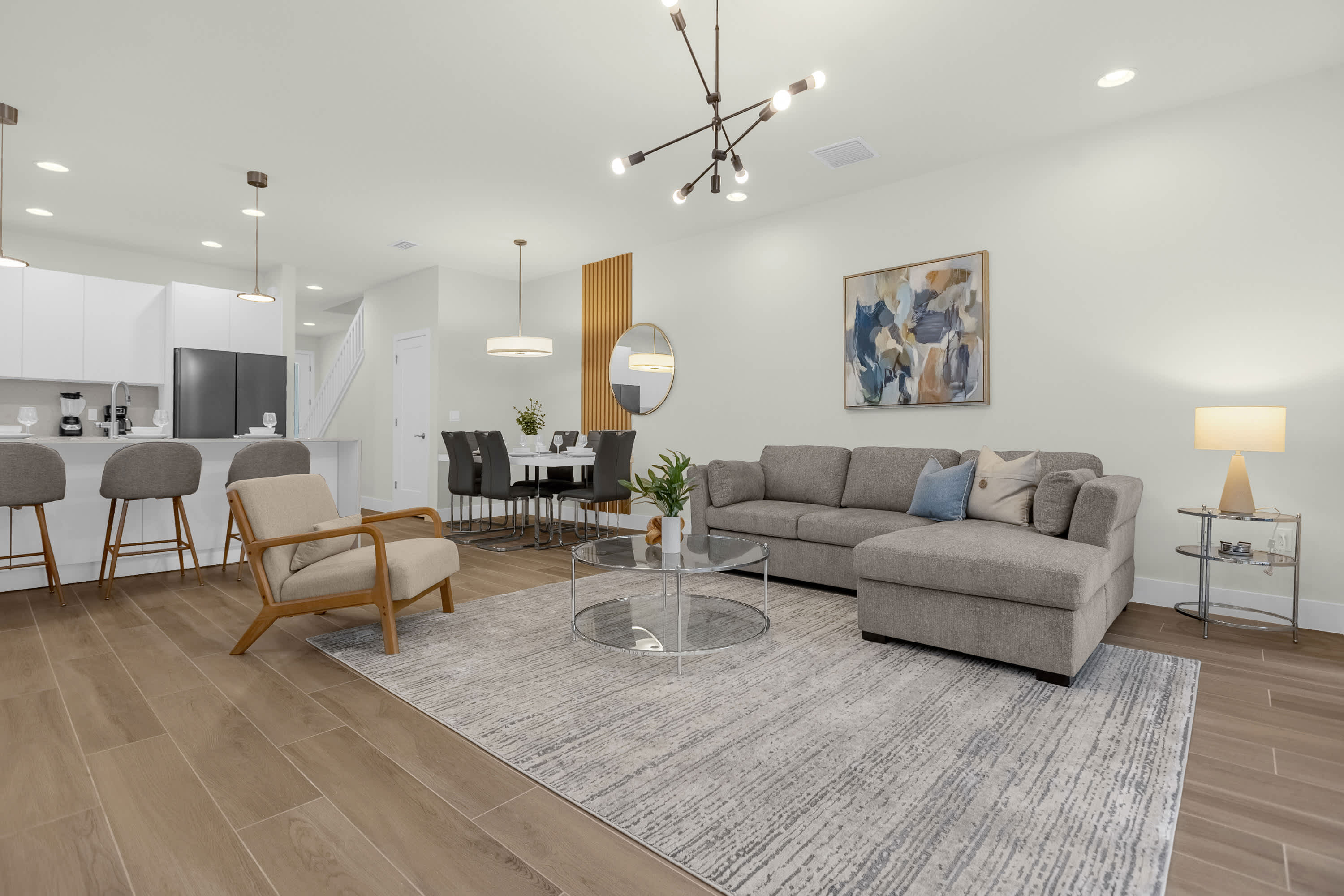 Wide view of the open-concept living area, showcasing a gray sectional sofa, a glass coffee table, and an accent chair on a large area rug. The dining area with a white table and the modern kitchen with white cabinetry are visible in the background.
