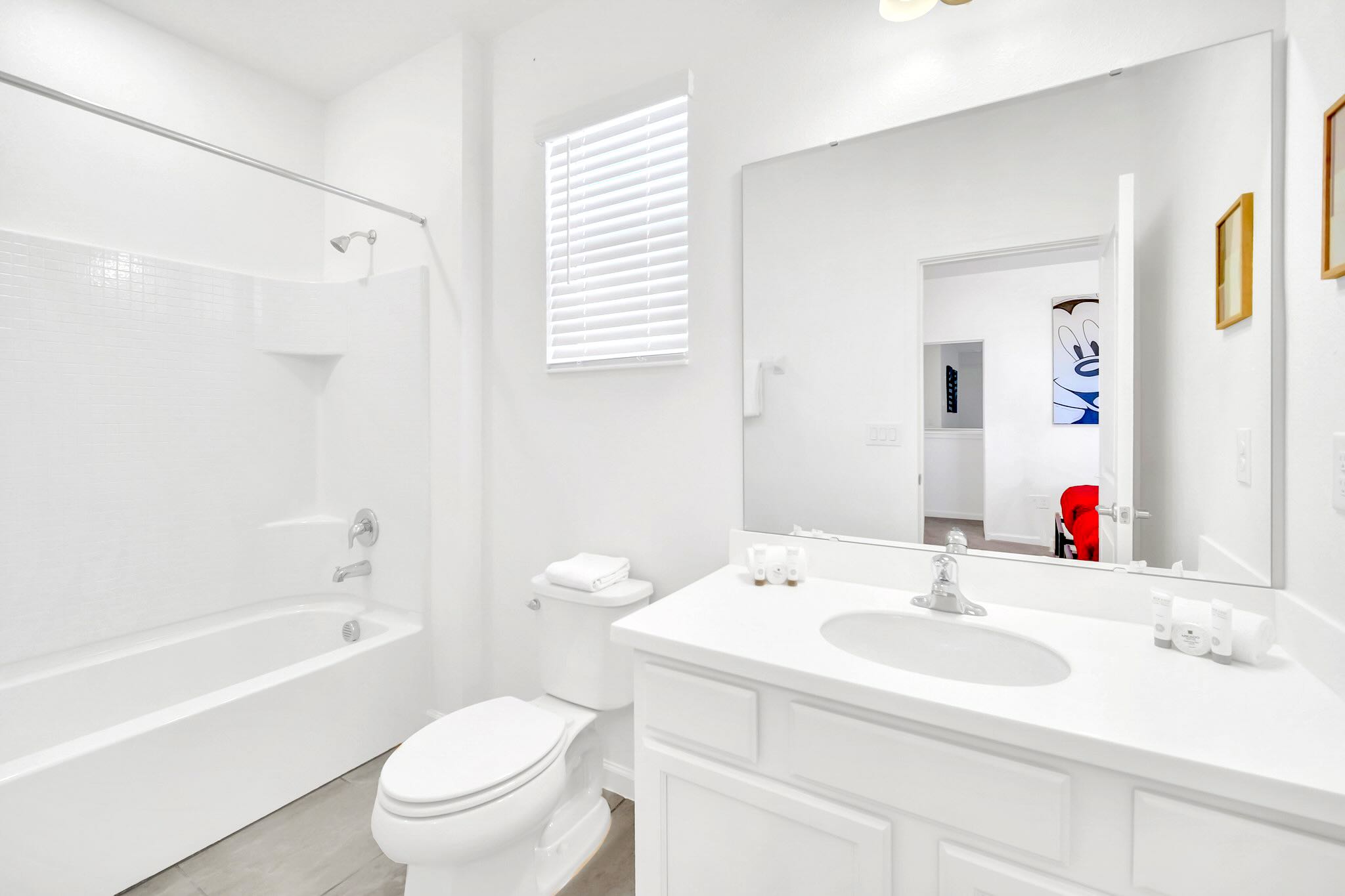 A pristine white bathroom featuring a single vanity with a large mirror, a toilet, and a shower/tub combination, with a window and blinds providing natural light.