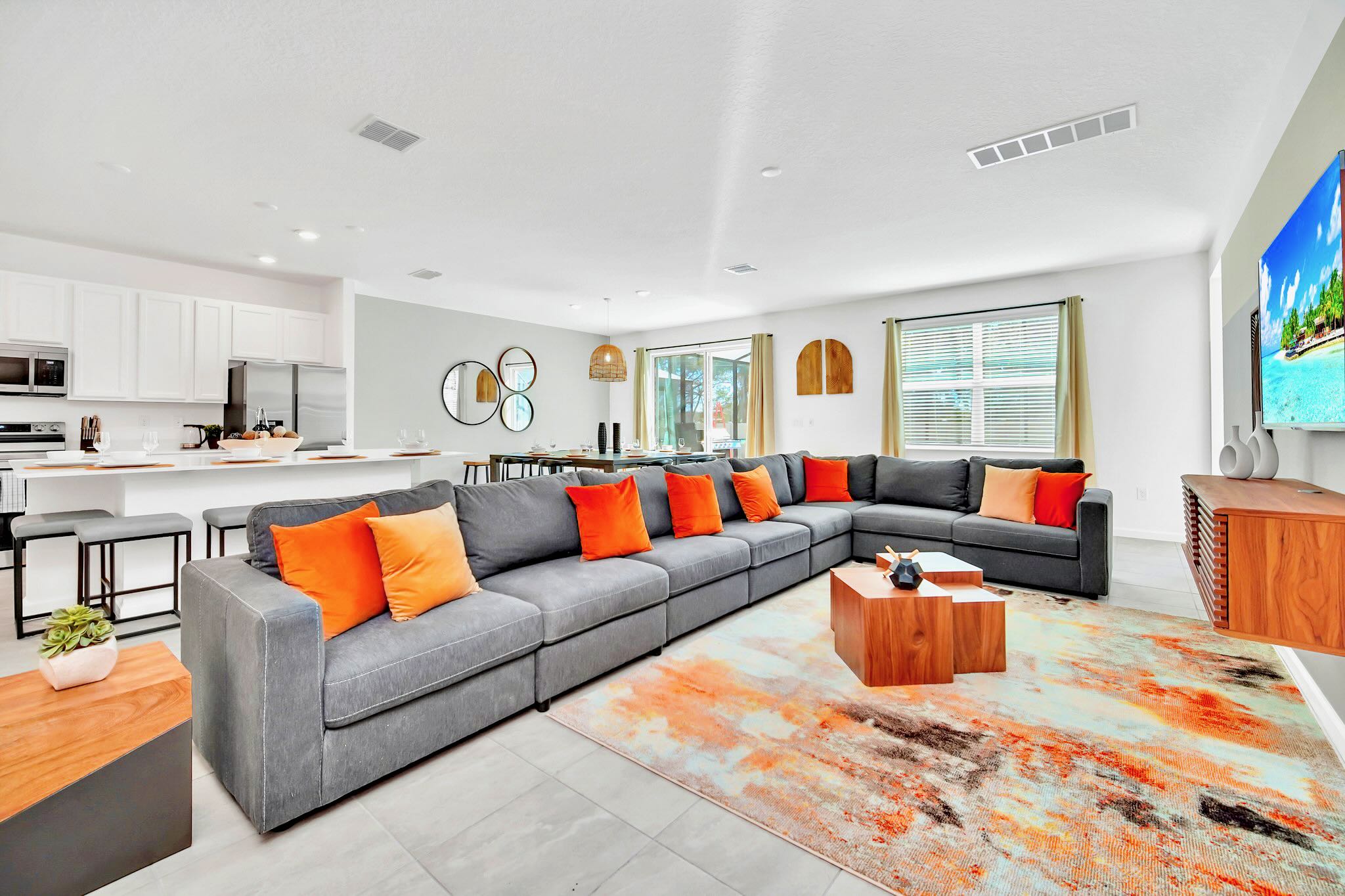 Bright open-concept living area featuring a large grey sectional sofa with vibrant orange throw pillows, a modern wooden coffee table, and a colorful abstract rug, flowing into a white kitchen and dining space.
