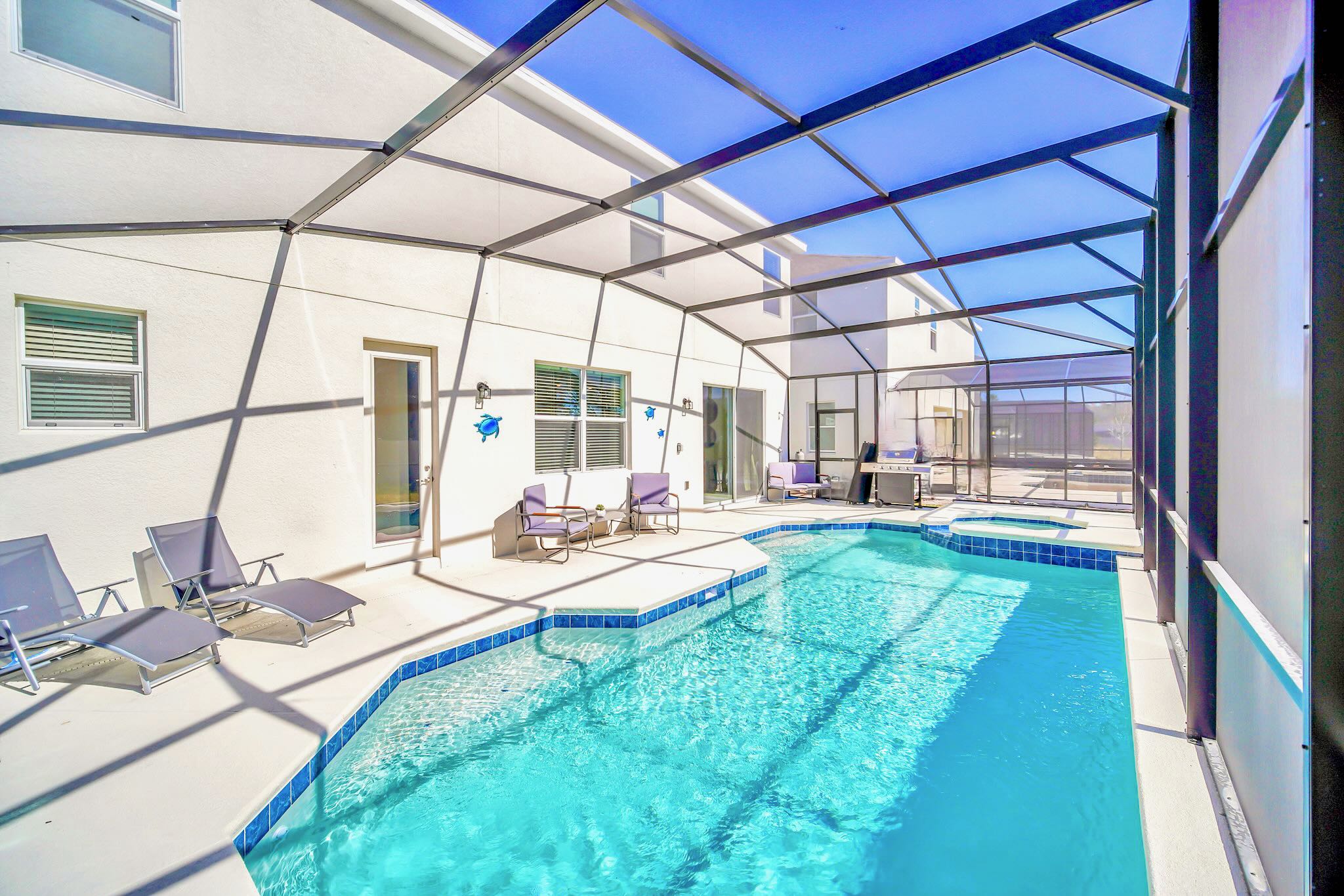 Wide view of the screened-in pool area, showcasing the swimming pool, hot tub, and multiple lounge chairs and seating areas on the concrete deck, with the white exterior of the house visible.