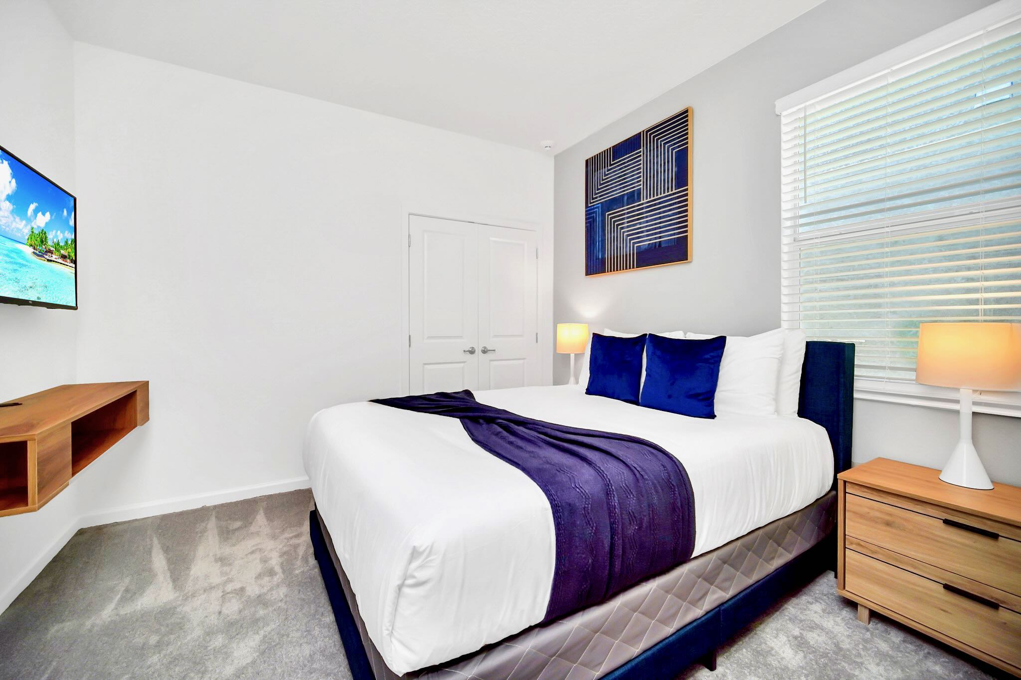 Modern bedroom featuring a queen bed with a dark blue upholstered headboard and matching bedding, light gray carpet, and a wall-mounted TV. A large abstract blue artwork hangs above the bed, and a wooden floating console is beneath the TV.