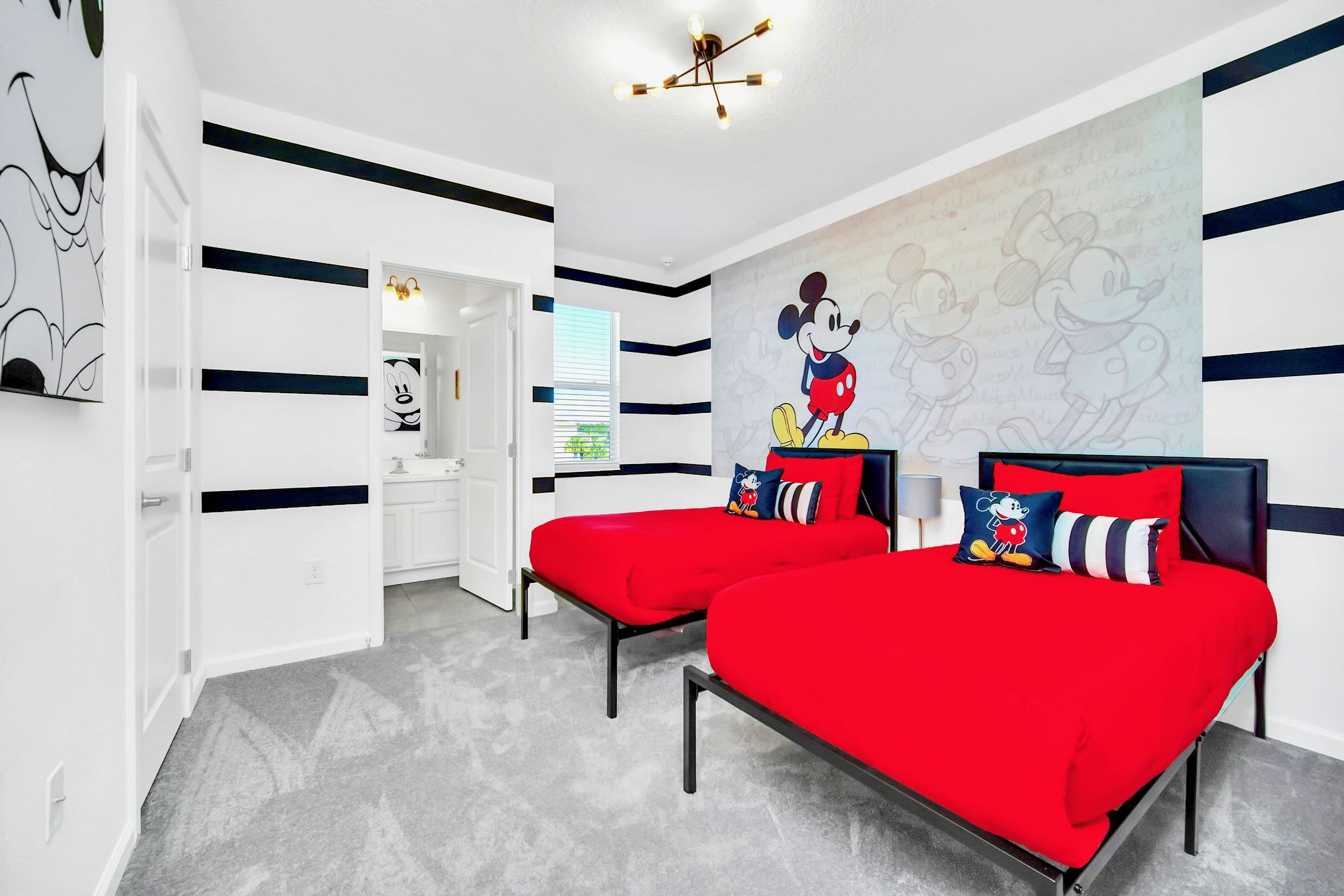 A vibrant Mickey Mouse themed bedroom with two twin beds featuring bright red bedding and character pillows, complemented by black and white striped walls and a large Mickey Mouse decal.