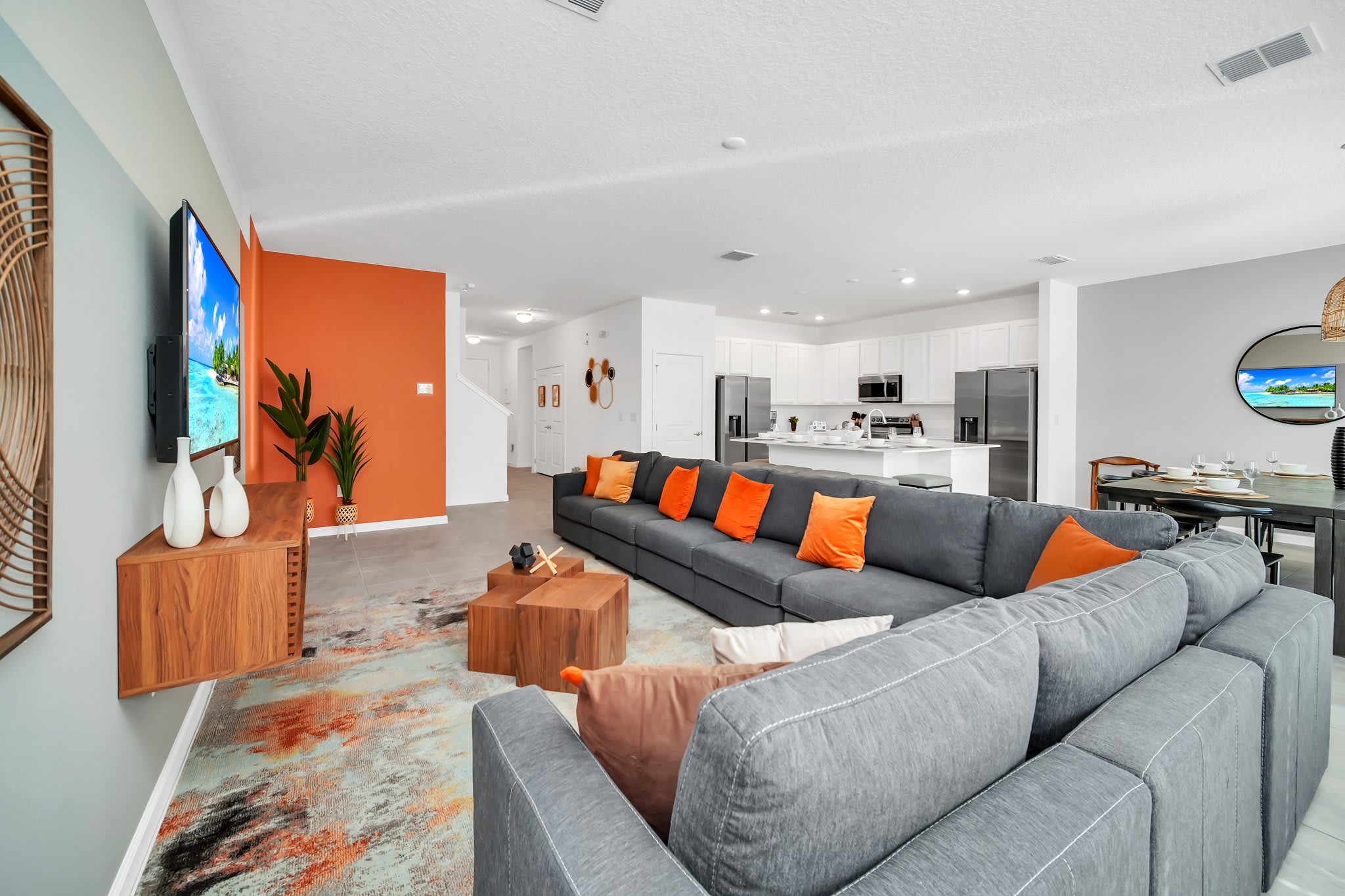 Spacious open-concept living room featuring a large gray sectional sofa with orange throw pillows, a vibrant abstract rug, and a wall-mounted TV above a wooden media console, with an orange accent wall.