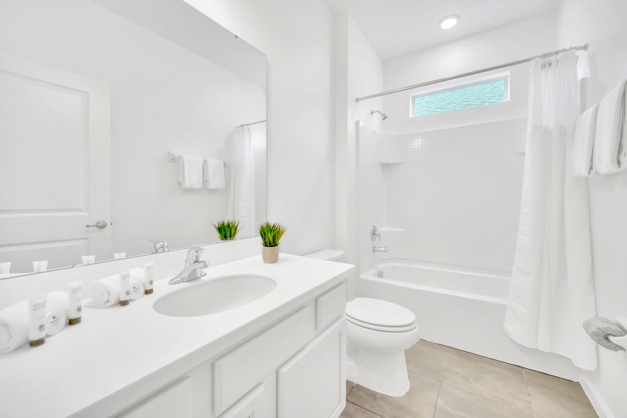 Bright, modern bathroom with a white single vanity, toilet, and a shower-tub combination. The room features light gray tiled floors, white walls, and a small window above the shower.