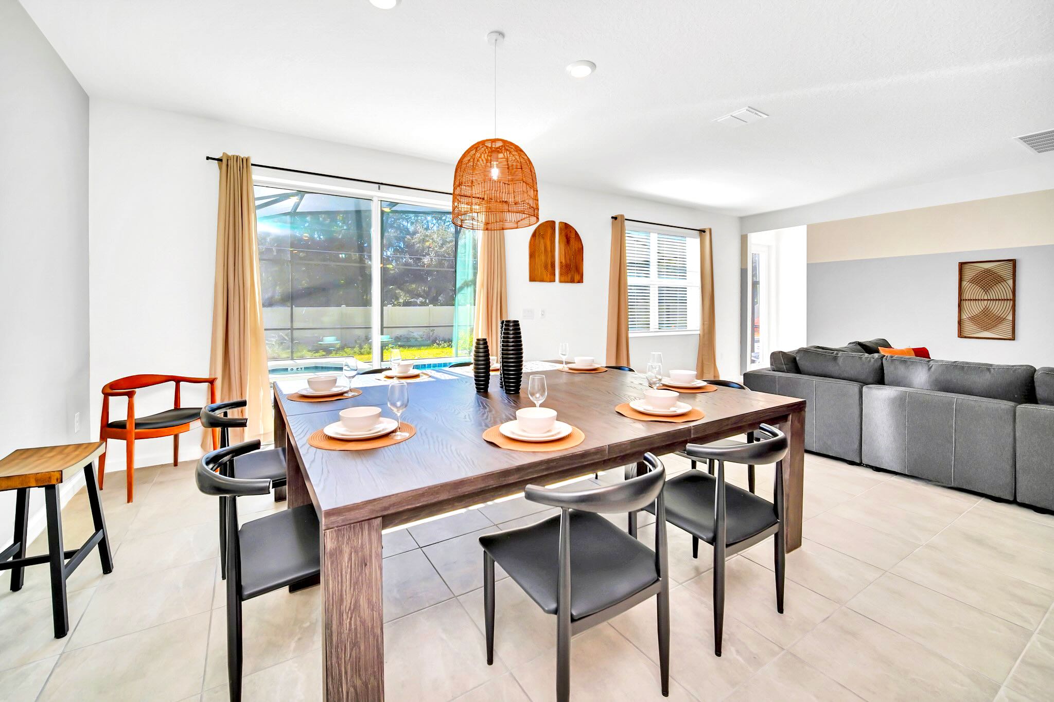 Modern dining area with a large dark wood rectangular table set for eight, black chairs, and a distinctive woven pendant light, with sliding glass doors leading to an outdoor pool area.