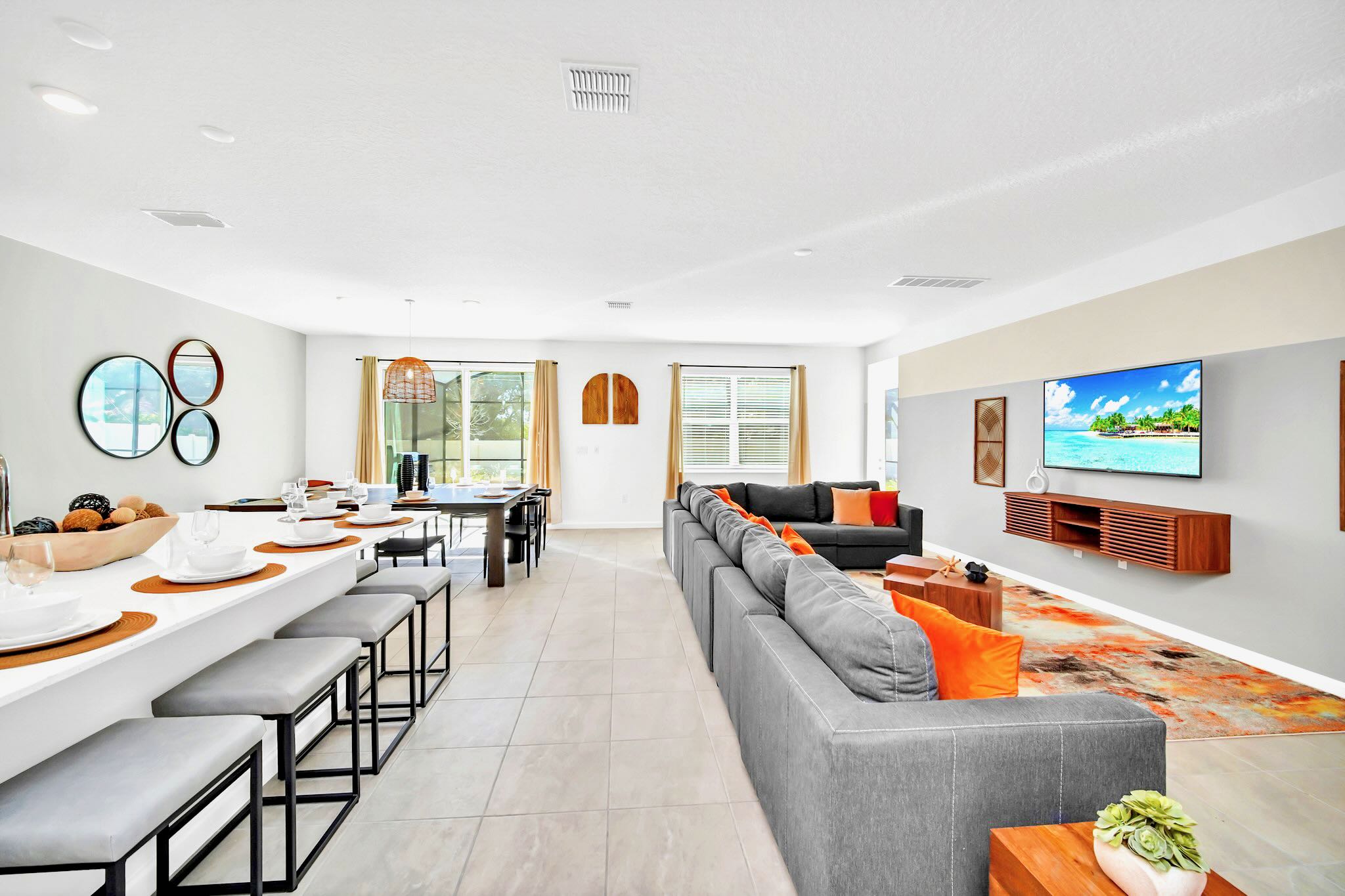 Wide view of the open-concept living space, showcasing a large gray sectional sofa, a wall-mounted TV, and extending into a dining area with a long table and a modern kitchen with a white island.