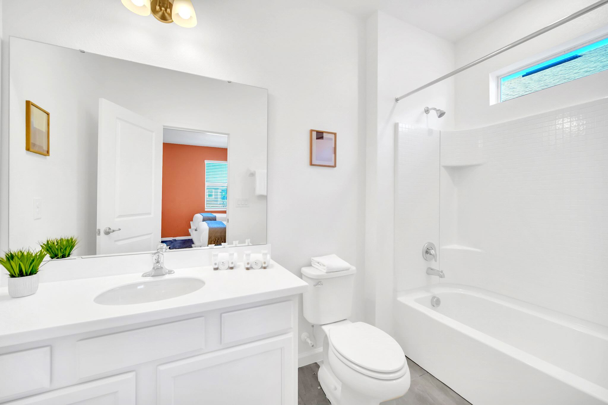 Clean bathroom with a white single vanity, toilet, and a shower-tub combination. The room has light gray tiled floors, white walls, and a small window above the shower, reflecting the orange bedroom.