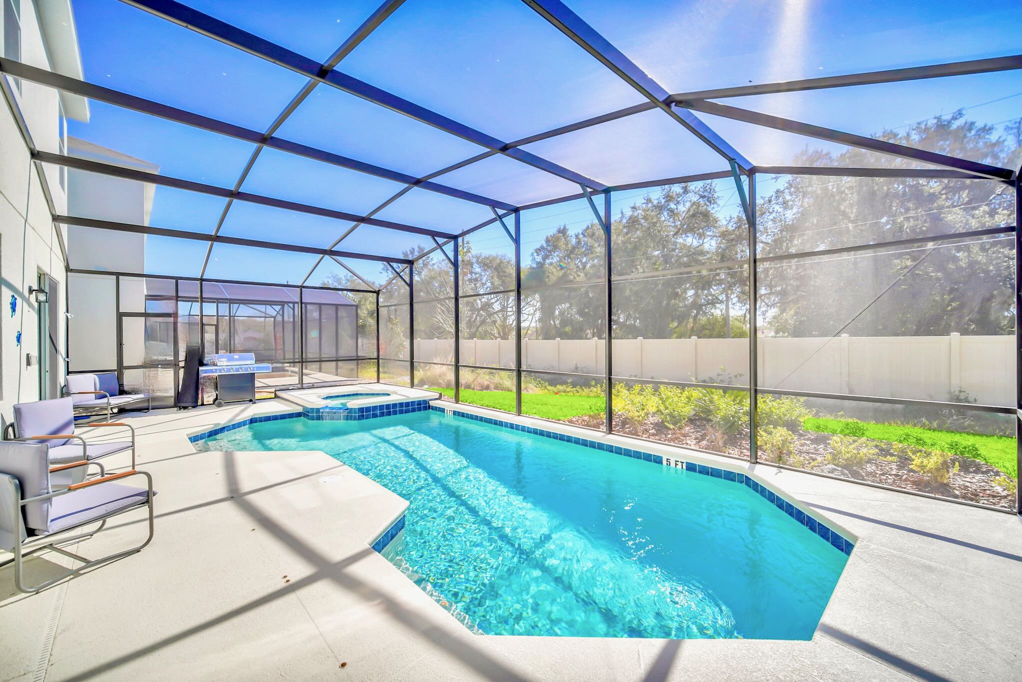Inviting screened-in outdoor pool area with a sparkling blue swimming pool and an adjacent hot tub, surrounded by a concrete deck and views of lush greenery beyond the enclosure.