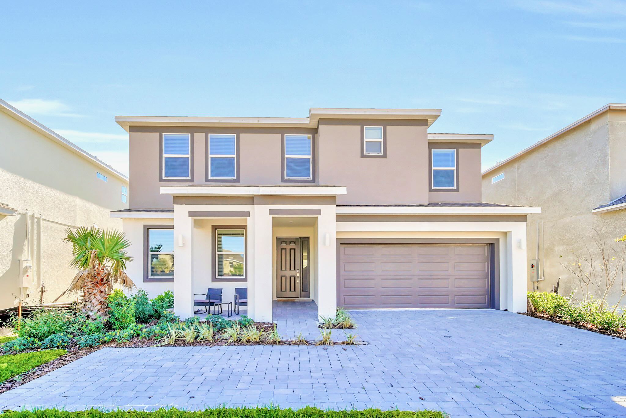 Modern two-story house exterior with a light brown and white facade, featuring a paved driveway leading to a two-car garage, a covered front porch with seating, and landscaped garden beds.