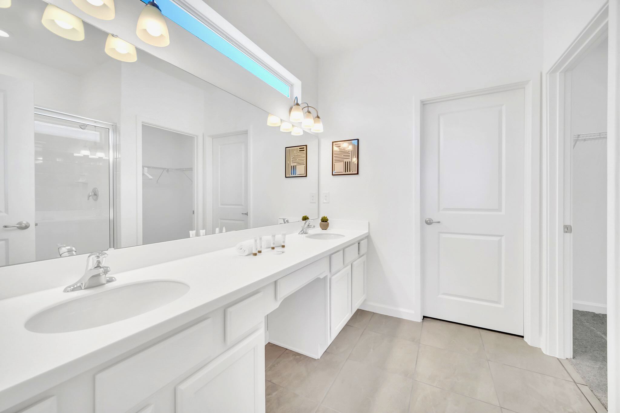 Bright, modern master bathroom featuring a long white double vanity with two sinks, a large mirror, and a glass-enclosed shower visible in the reflection. The room has light gray tiled floors and white walls.