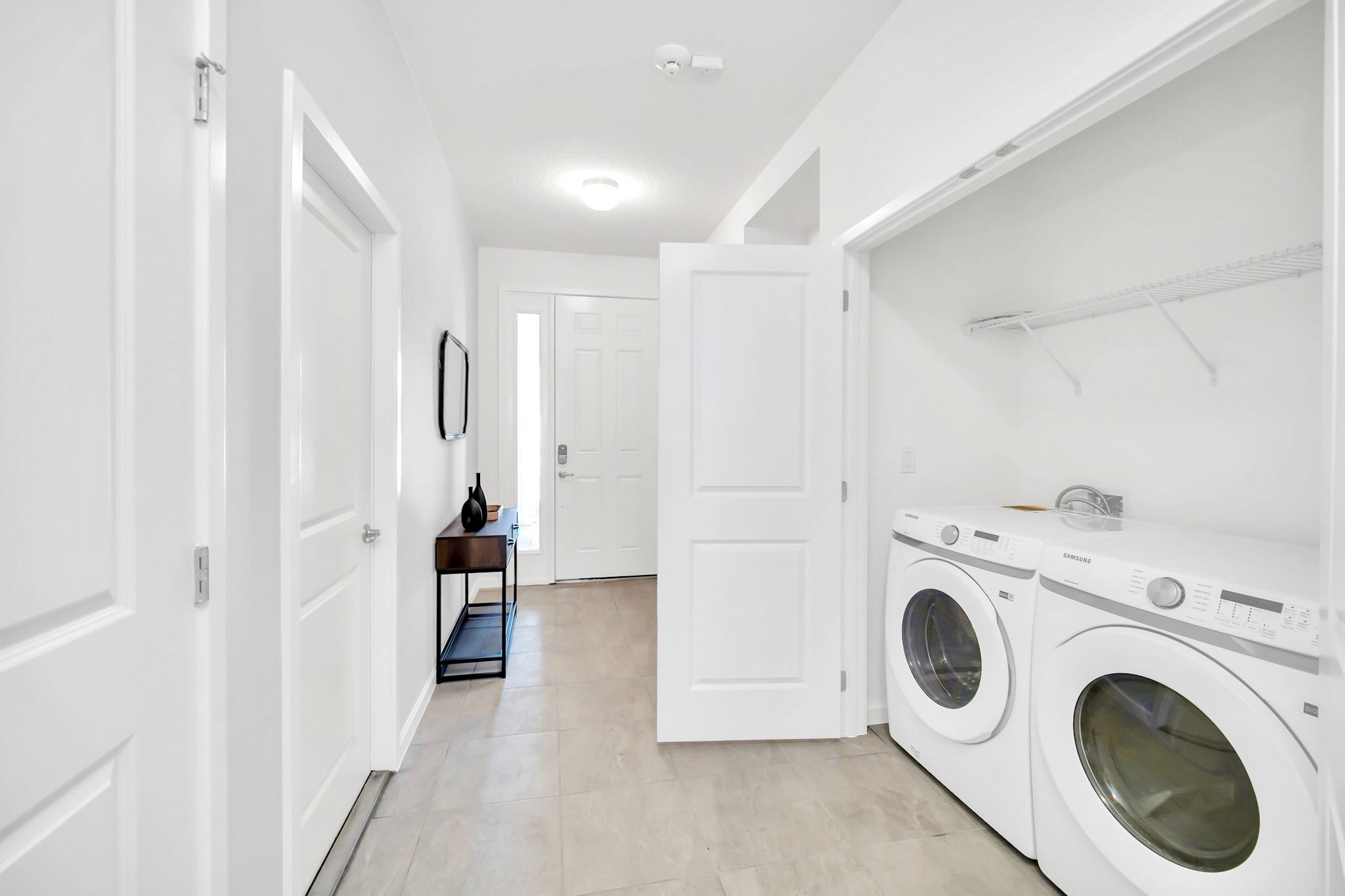 A bright hallway leading to the front door, featuring a discreet laundry closet with a front-loading washer and dryer, and a small console table with decorative items.