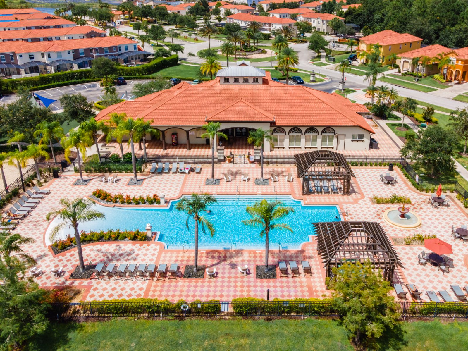 An aerial view of a large communal swimming pool area, featuring a clubhouse, numerous lounge chairs, lush palm trees, and decorative patterned paving.