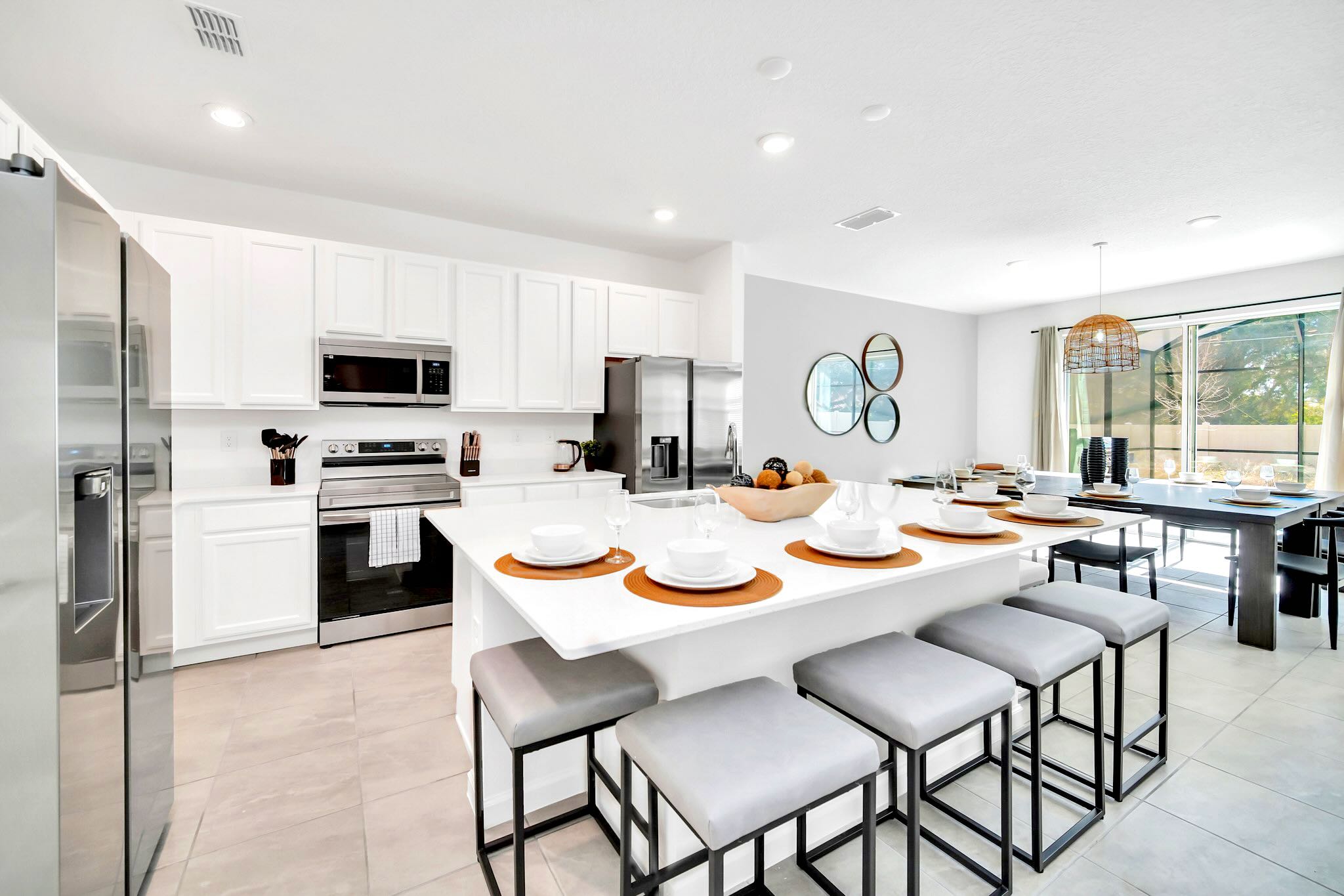 Modern white kitchen with stainless steel appliances, a large island featuring bar stool seating, and an adjacent dining area with a dark wood table, all set on light tile flooring.