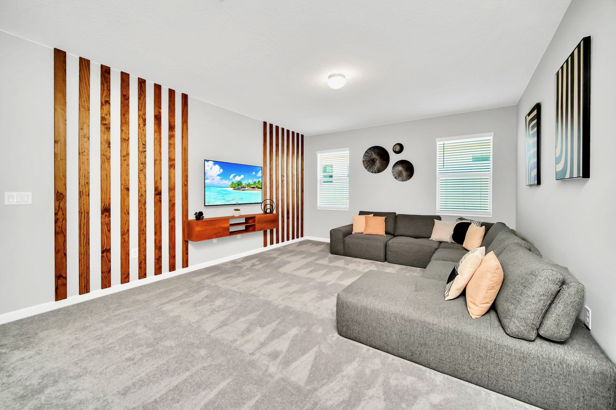 Upstairs game room or secondary living area with a large gray sectional sofa, a wall-mounted TV against a decorative wooden slat wall, and light gray carpeted flooring.