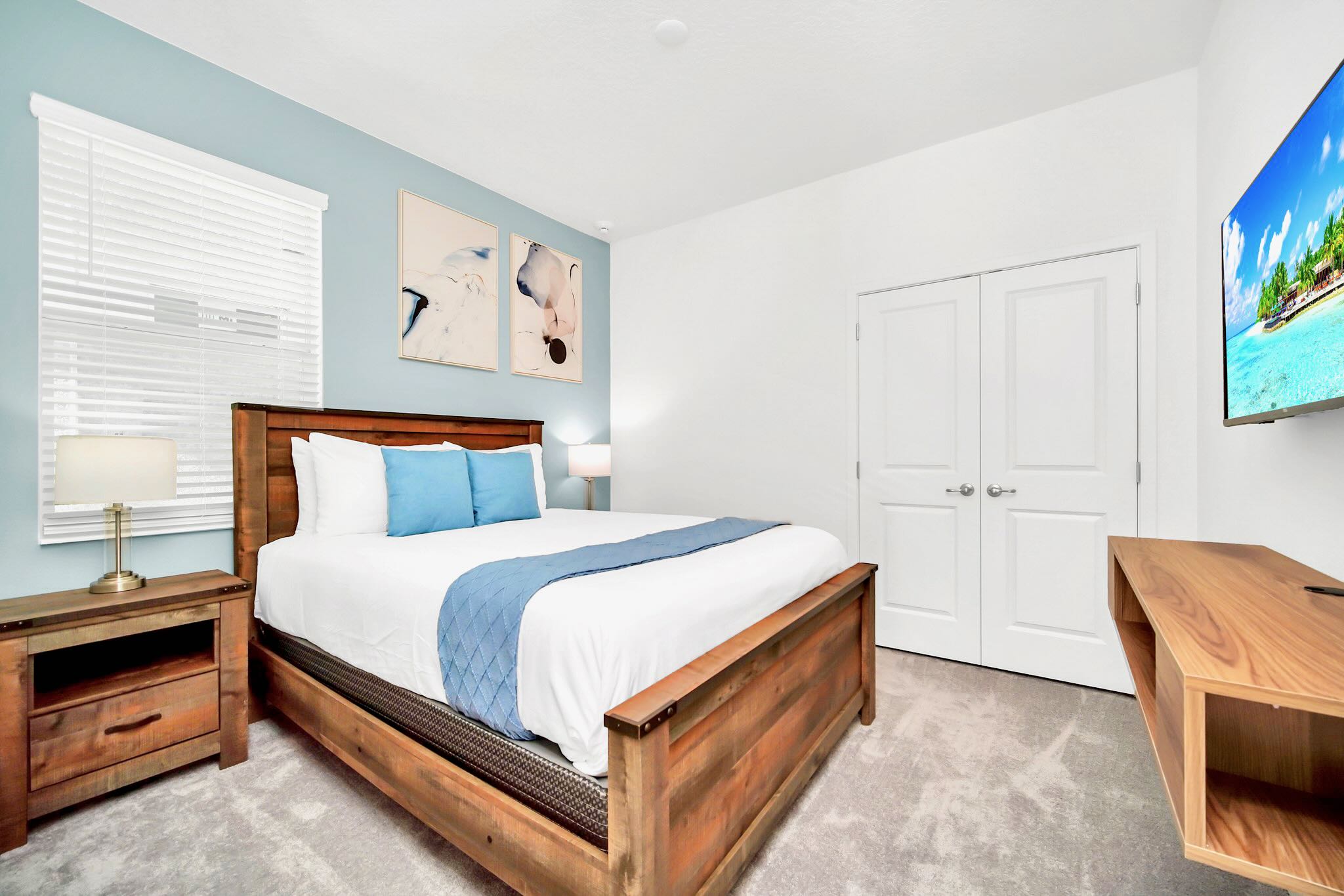 Cozy bedroom with a rustic wooden queen bed, light blue accent wall, and a wall-mounted TV. The room features light gray carpet, white blinds, and two framed abstract artworks above the bed.
