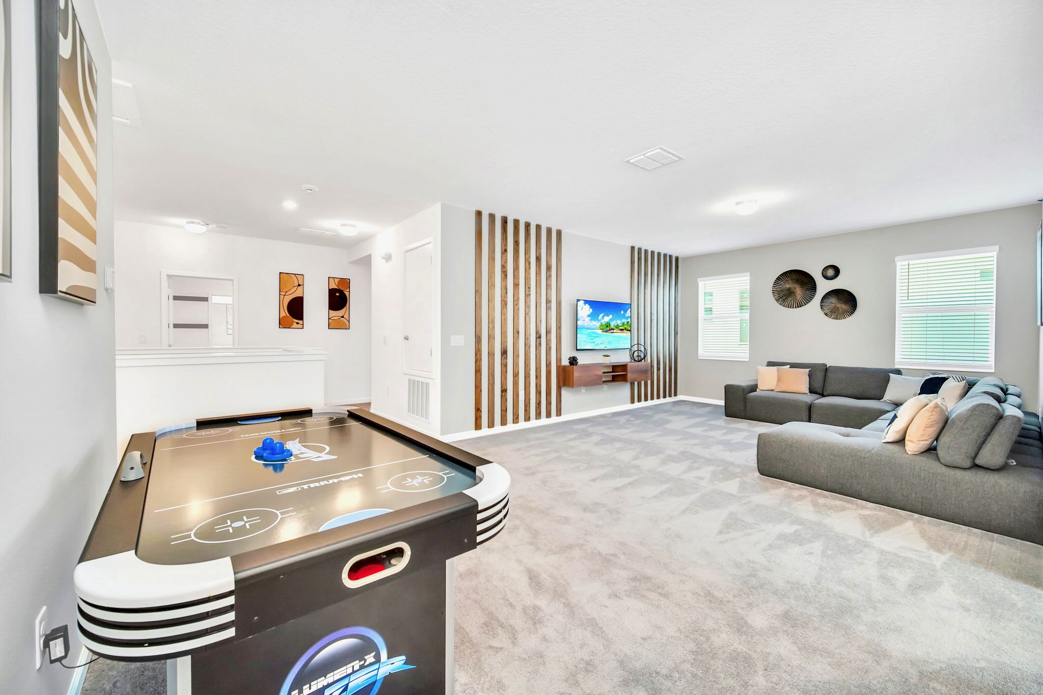 Spacious upstairs loft area featuring an air hockey table and a grey sectional sofa facing a wall-mounted TV, set on light grey carpet with modern wooden slat wall decor.