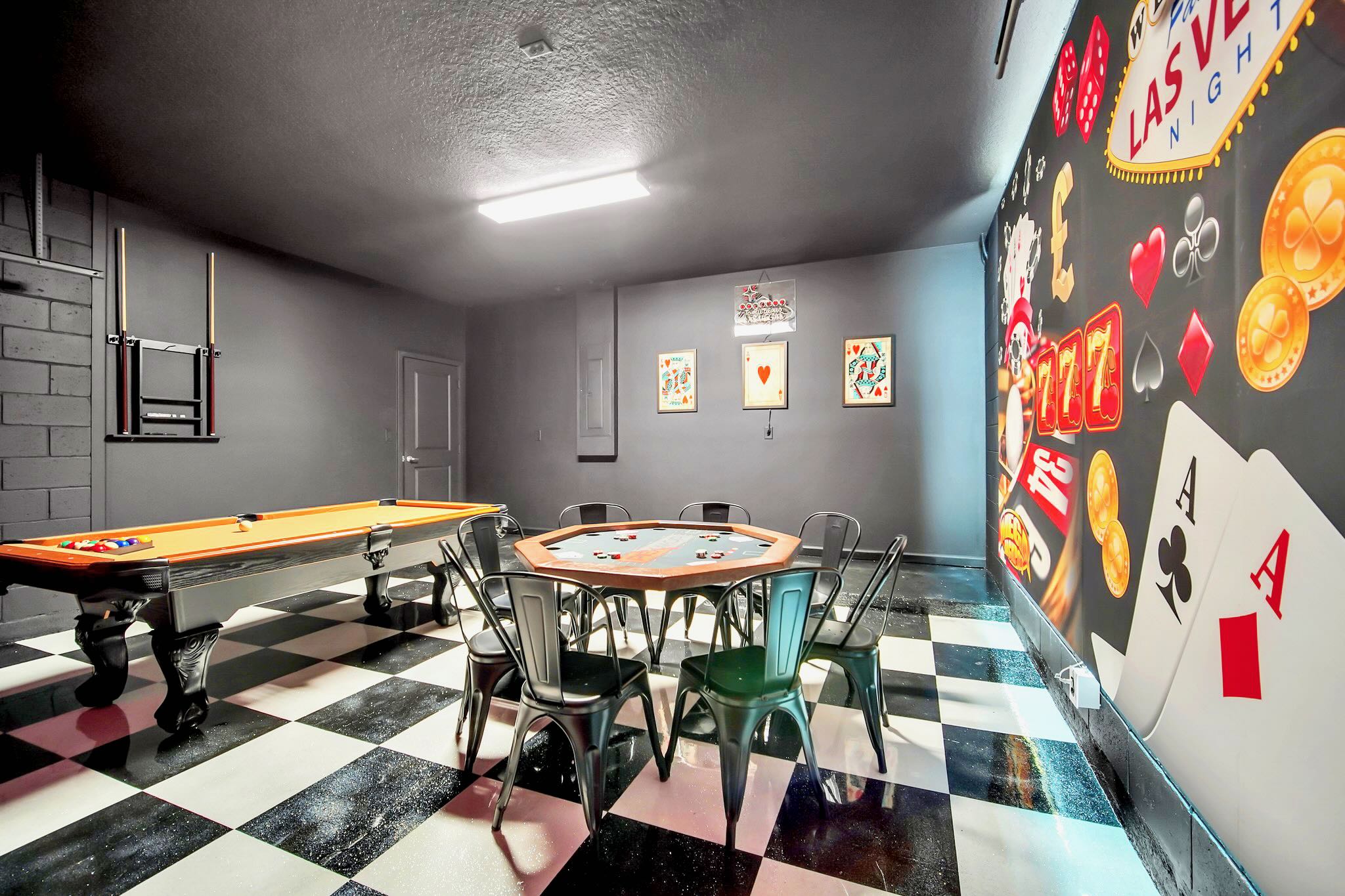 Alternative view of the game room with a black and white checkered floor, highlighting an octagonal poker table with chairs, a pool table, and casino-themed wall art.