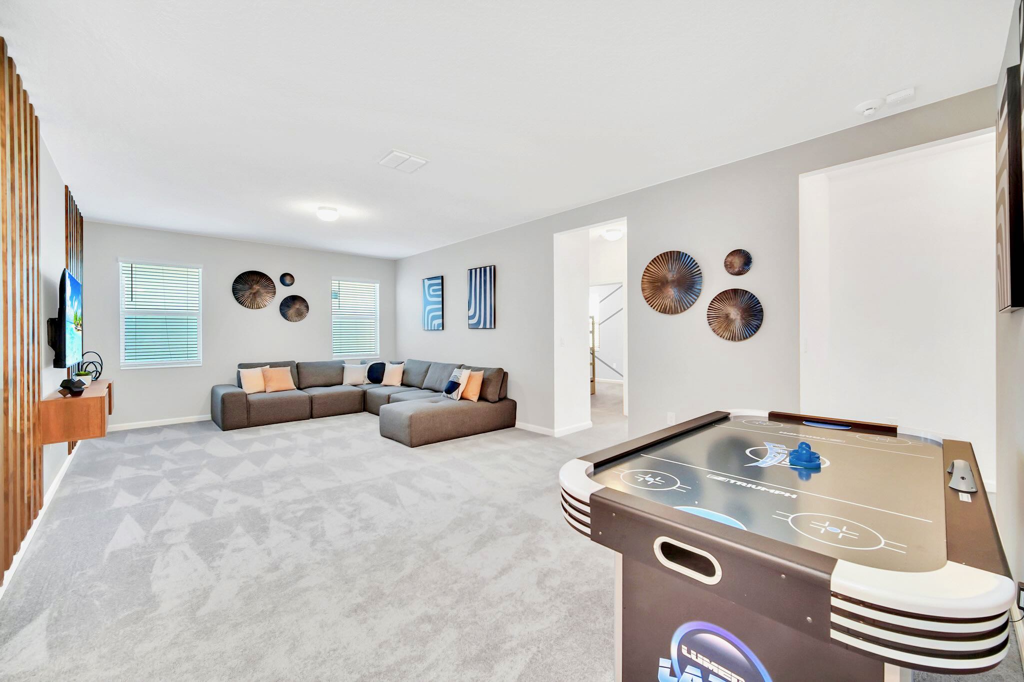 Spacious game room featuring an air hockey table, a comfortable gray sectional sofa, and a wall-mounted TV set against a stylish wooden slat accent wall, all on a light gray carpet.